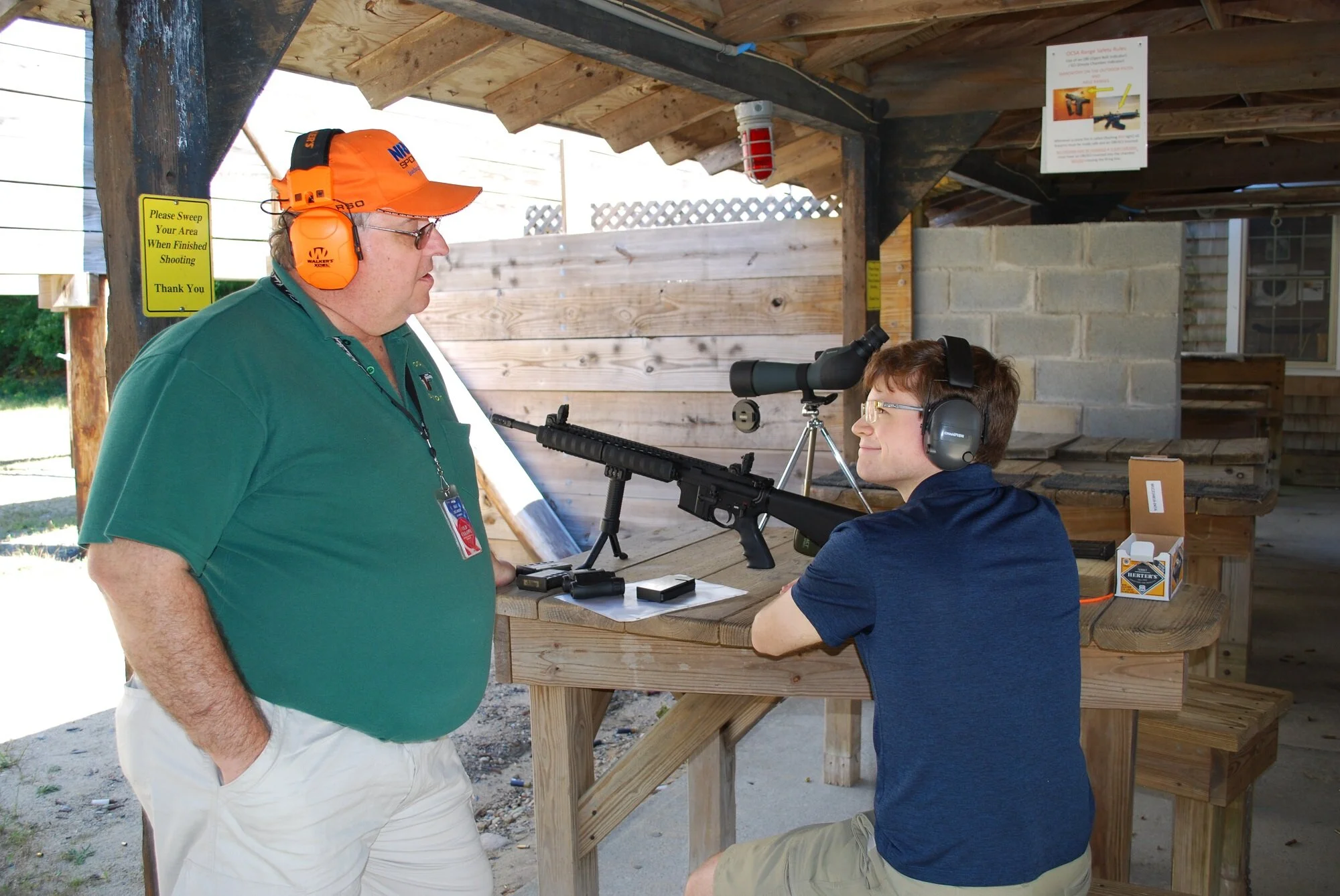 A young man with glasses and ear protection sitting at a shooting range, aiming a rifle with a scope, as an older man in a green polo shirt and orange hearing protection talks to him. The range is outdoors with a wooden structure and targets.