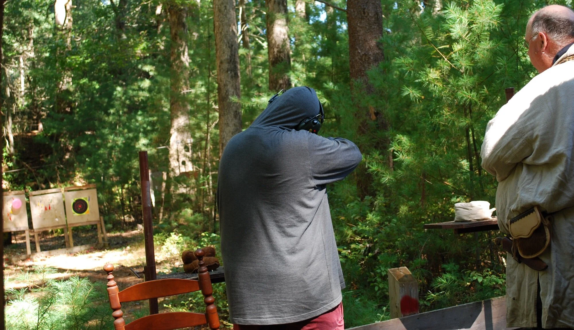 Two people at a shooting range in a forest, one wearing a hoodie and ear protection aiming a firearm, and the other wearing a beige jacket observing with arms crossed, with target boards in the background.