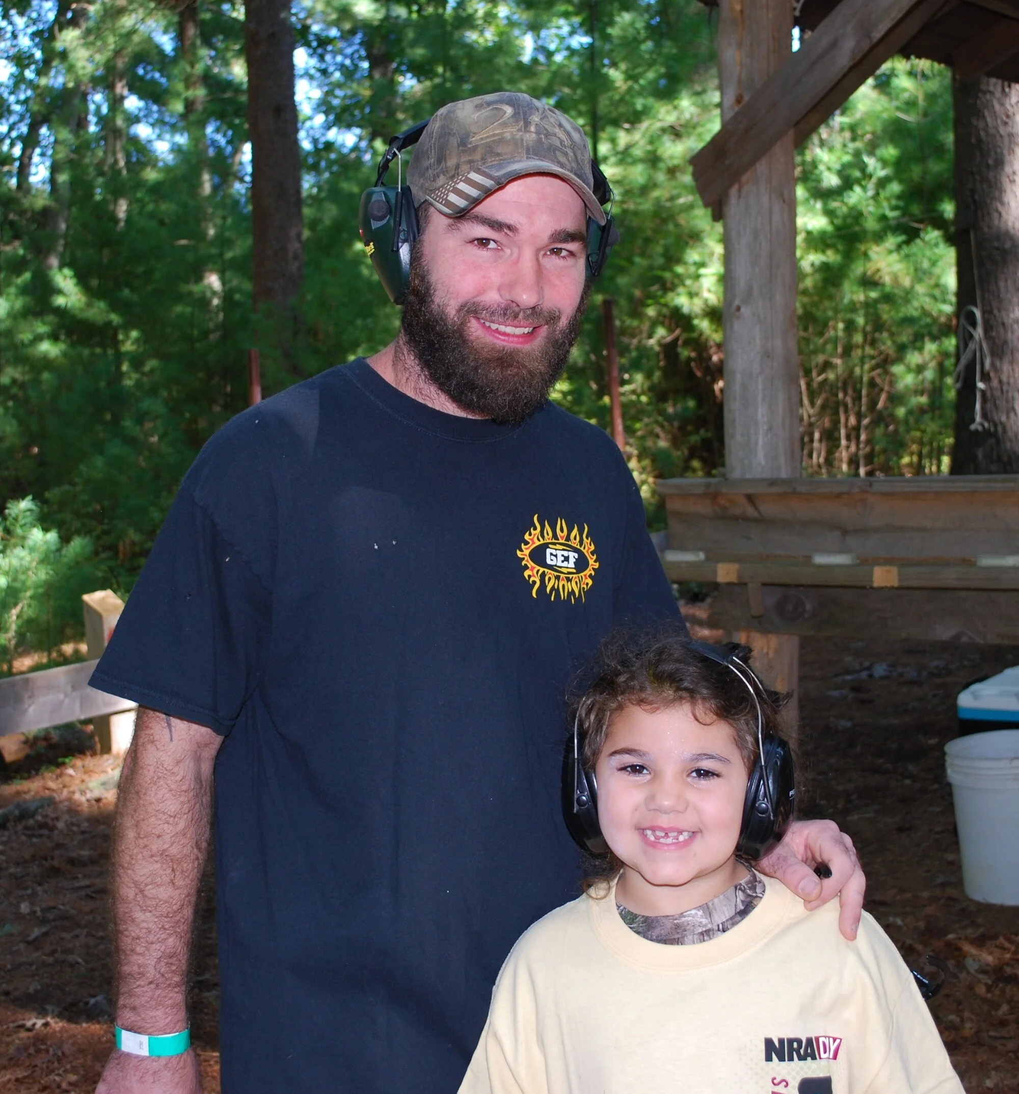 A man and a young girl smiling outdoors at a wooded area, both wearing protective hearing headphones. The man has a beard, is wearing a black T-shirt with a logo, and a camouflage baseball cap. The girl has curly hair and is wearing a yellow T-shirt 