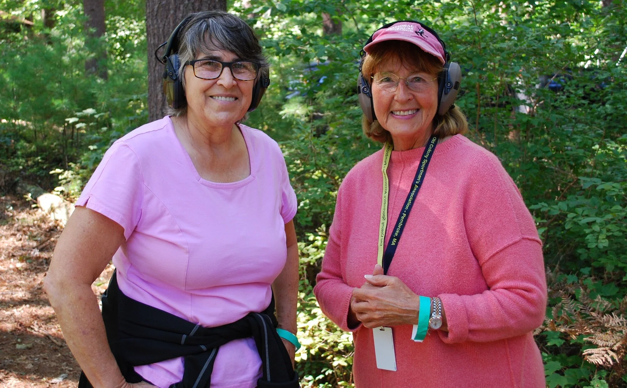 Two smiling women in pink shirts and glasses wearing headphones, standing outdoors in a wooded area.