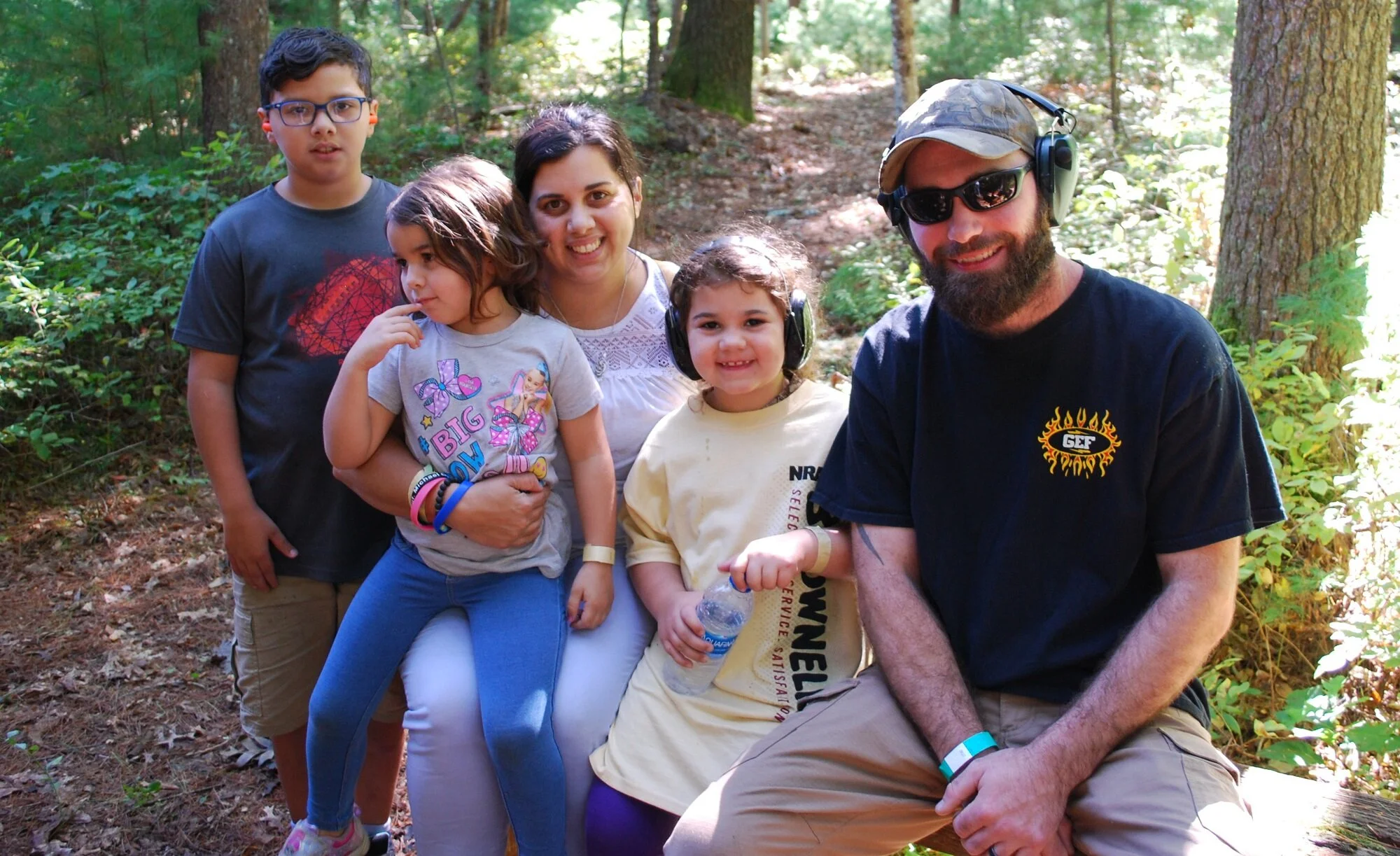 A group of six people, including three children, a woman, and two men, sitting and standing in a forested area with trees and sunlight.