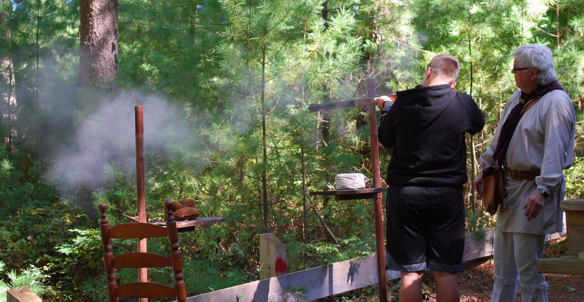 Two men are in a forest, with one aiming a rifle at a target on a shooting stand while the other watches. The man aiming the rifle is wearing a black hoodie, and the other man has white hair, glasses, and is dressed in a light-colored striped shirt a