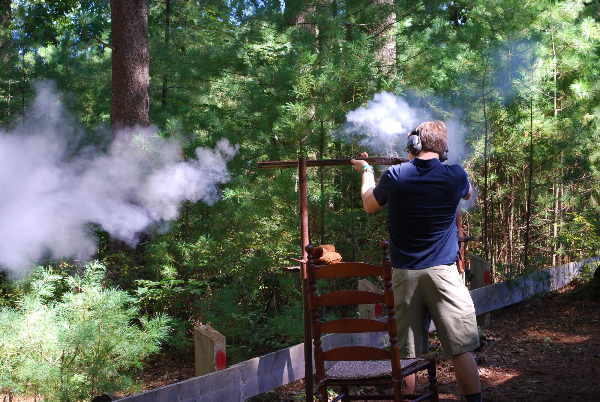 A man wearing headphones and dark blue shirt aiming a long rifle in a wooded outdoor setting, with smoke around the barrel.