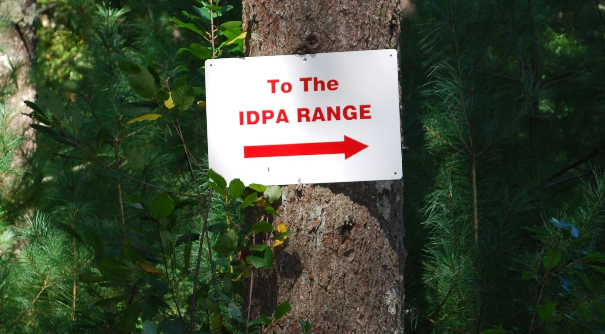 A white sign with red text and arrow, attached to a tree, directs to the IDPA Range through a wooded area.