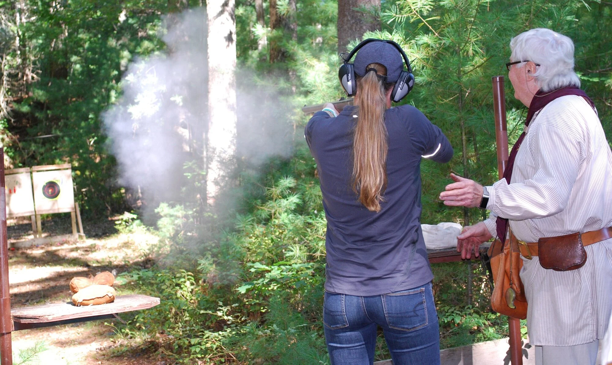 A woman firing a handgun at a shooting range outdoors, with another woman coaching or instructing. The woman shooting has long hair, headphones, a cap, and jeans, while the instructor has white hair, glasses, and a striped shirt.