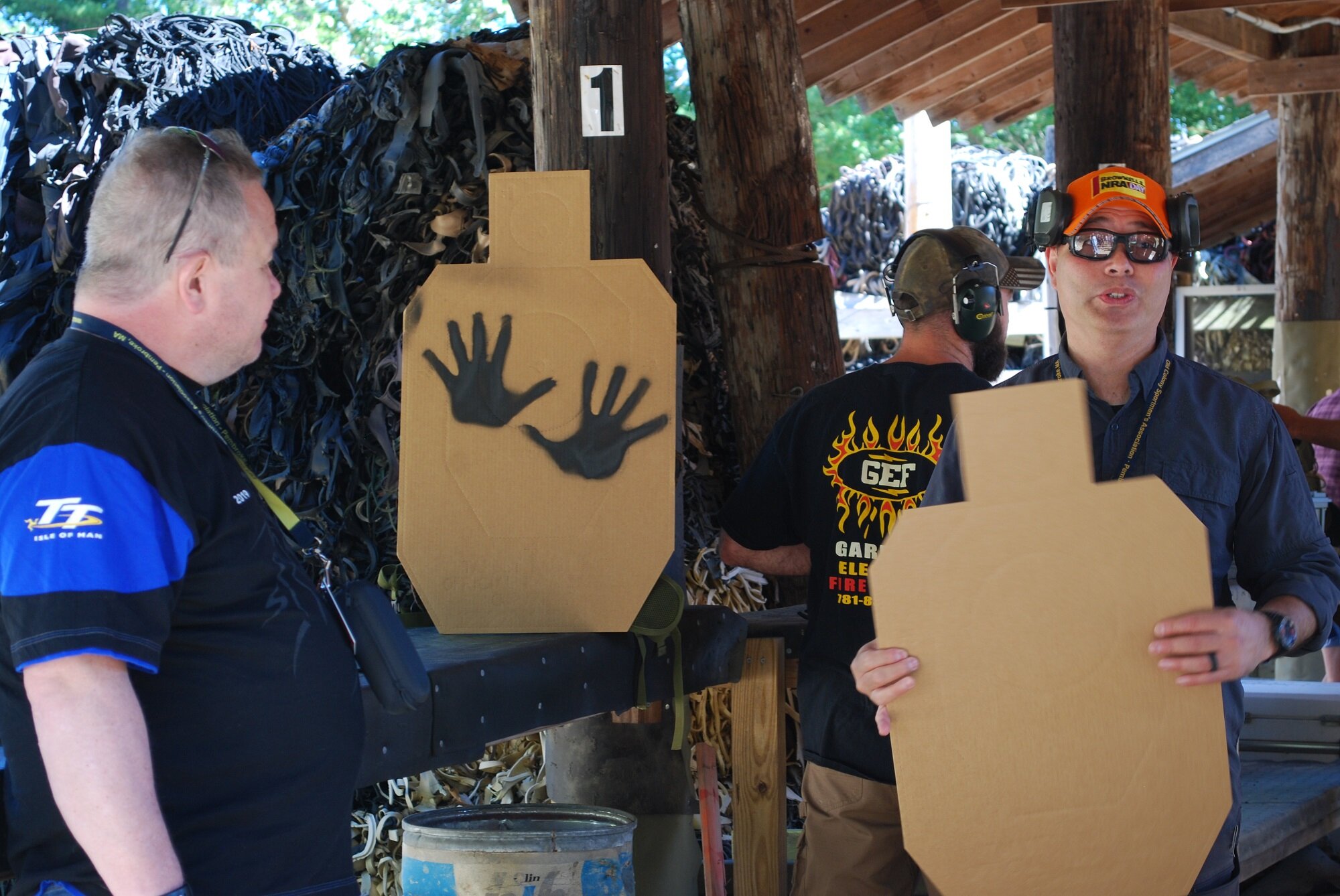 Group of men participating in a shooting event, wearing protective ear and eye gear, with cardboard target cutouts and a backdrop of discarded rubber.