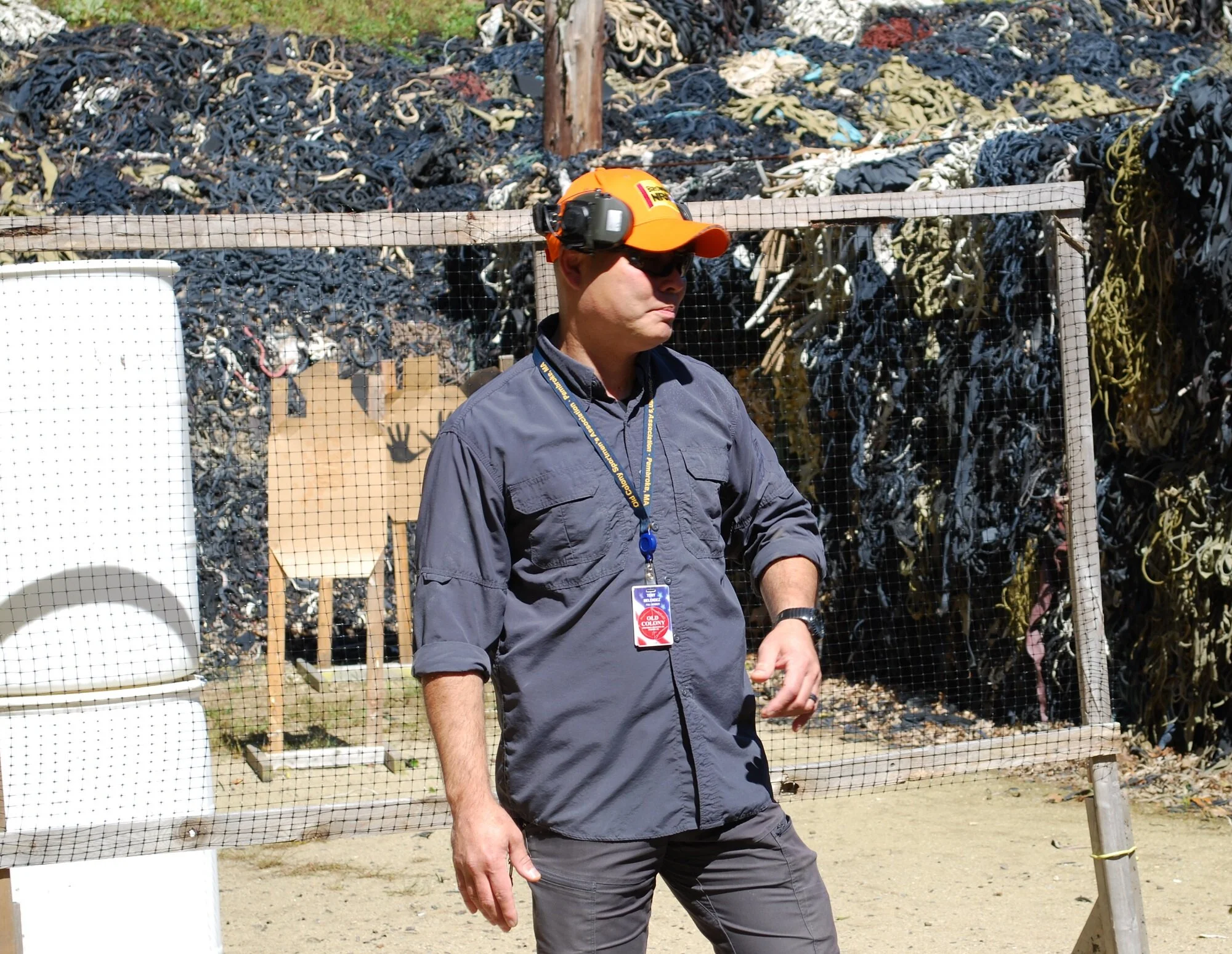 Man wearing an orange cap, sunglasses, and a gray long-sleeve shirt stands outdoors with a backdrop of stacked bags of tangled cords or ropes, a white object resembling a toilet, and a cardboard target with a silhouette of a hand, all behind a wire f