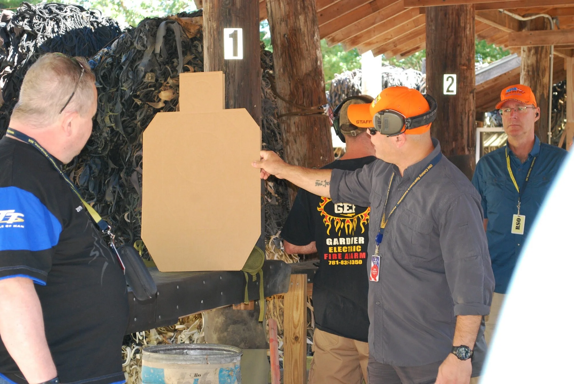 Group of men inspecting a shooting target at an outdoor shooting range. One man is pointing at the target, which is a cardboard cutout shaped like a gun target, with a pile of used gun magazines in the background.