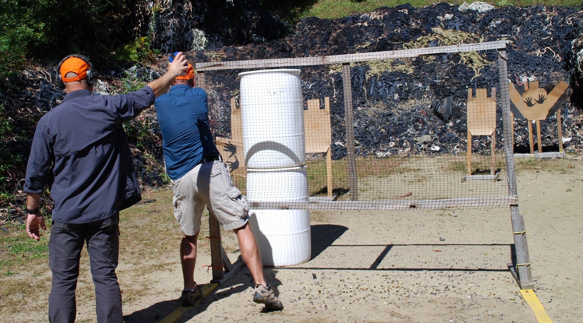 Two men at a shooting range, one is instructing the other. The shooting range has targets made from cardboard and a large barrel used as cover. The background shows a rocky, blackened area.