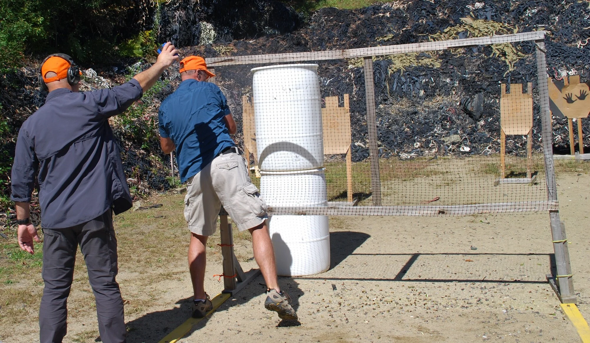 Two men, one with a gray jacket and orange cap and the other with a blue shirt and beige shorts, at a shooting range with targets and cardboard cutouts, preparing to shoot.