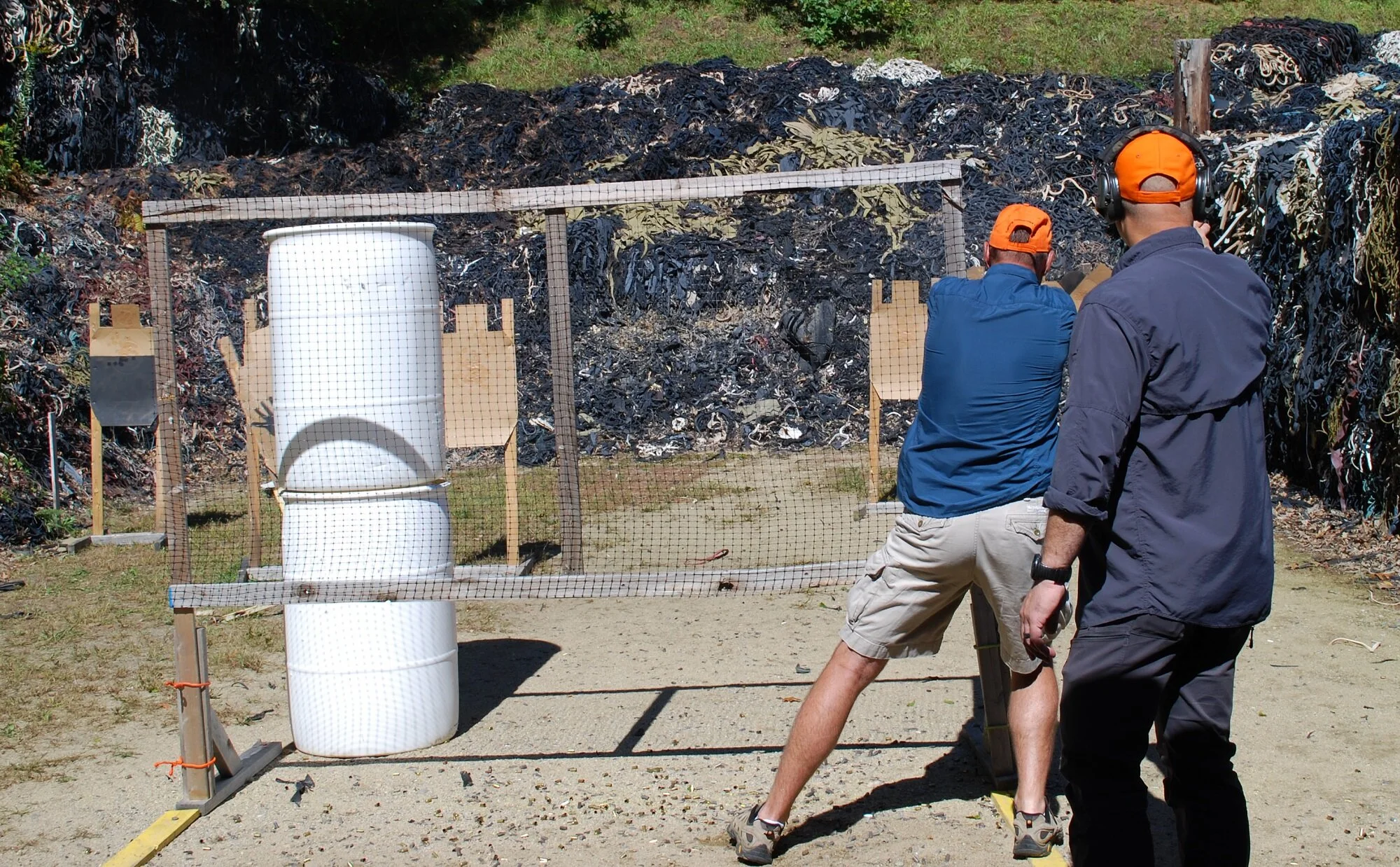 Two people shooting at a paper target on a shooting range, with a barricade and a plastic barrel behind them, and tall debris piles in the background.