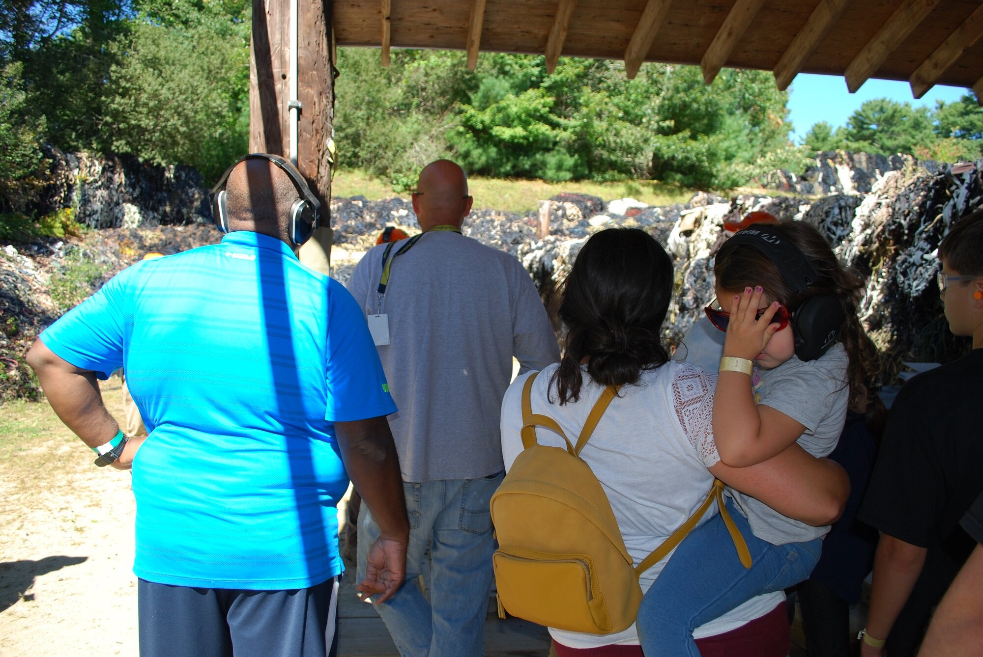 Group of people at an outdoor shooting range, wearing headphones and safety glasses, preparing to shoot at targets in a wooded area.