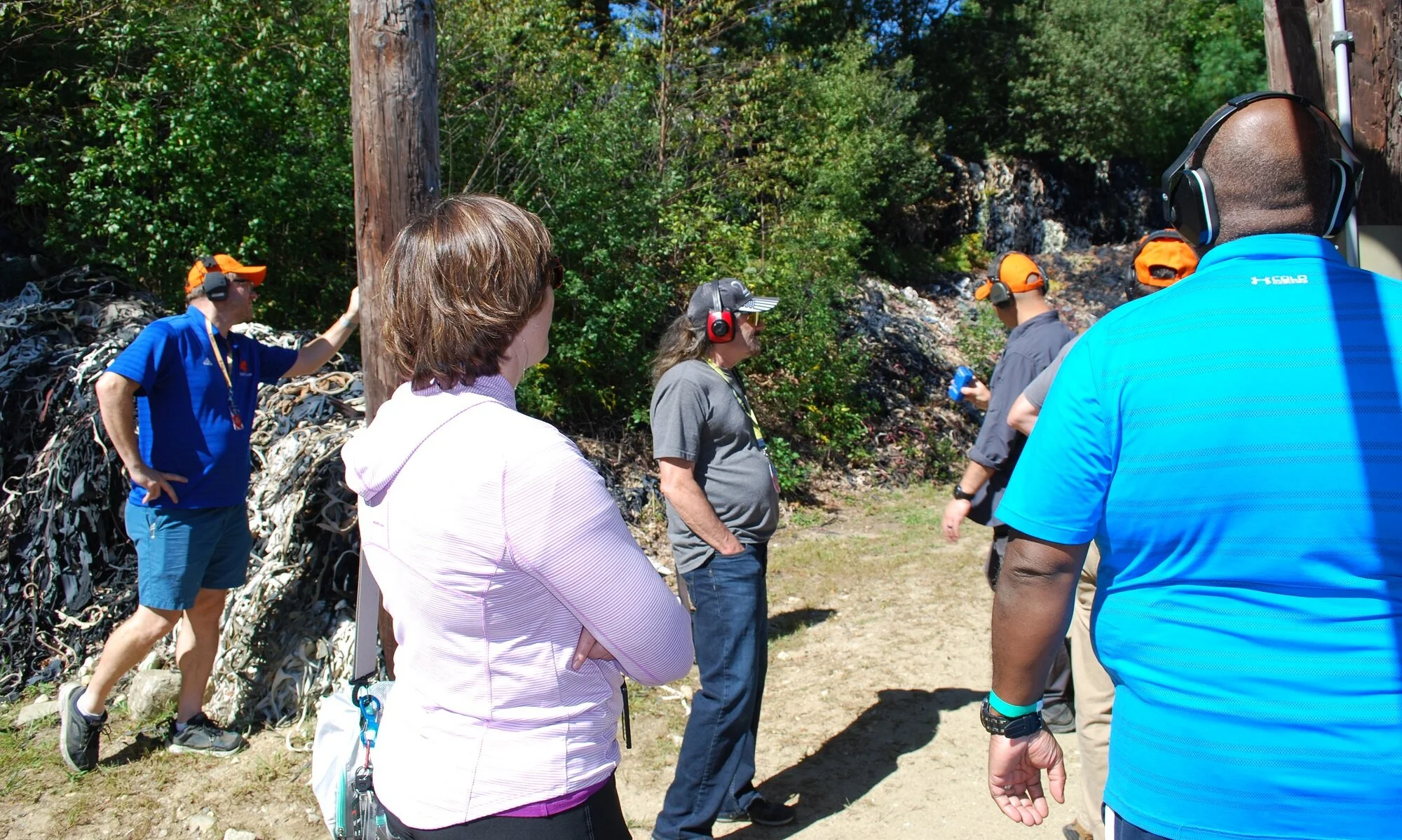 Group of people wearing headphones outdoors, standing near trees with some looking towards a speaker and others engaging in conversation.