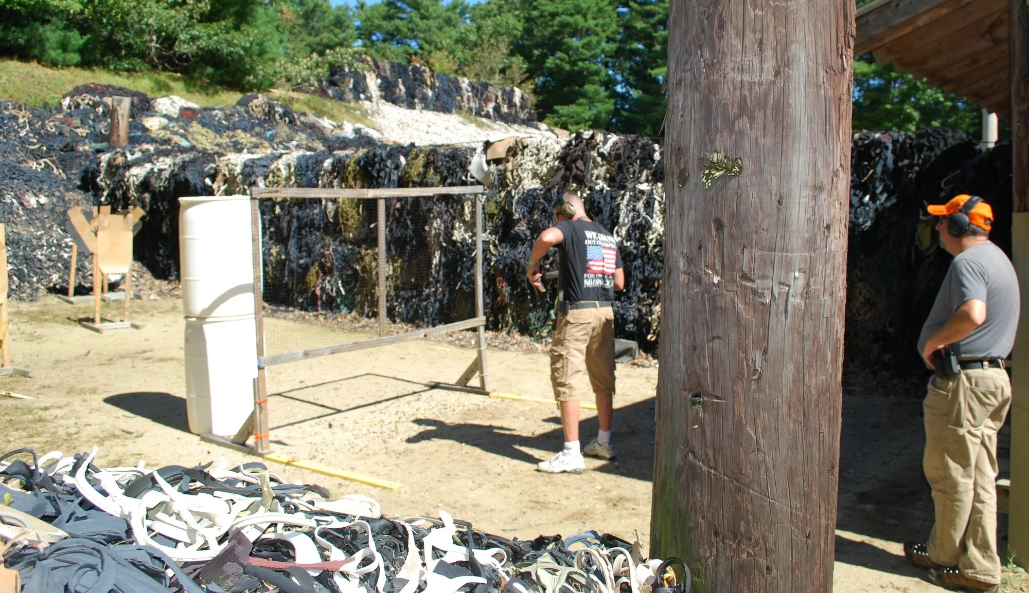 Two individuals at a shooting range, one adjusting rifle targets and another watching, with a large wooden utility pole, and a rocky backdrop with trees.