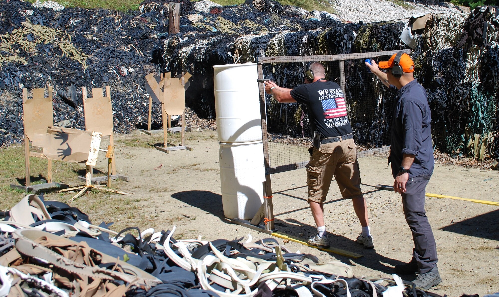 Two men at a shooting range, one aiming a handgun at targets, with a large pile of rubber on the backdrop.