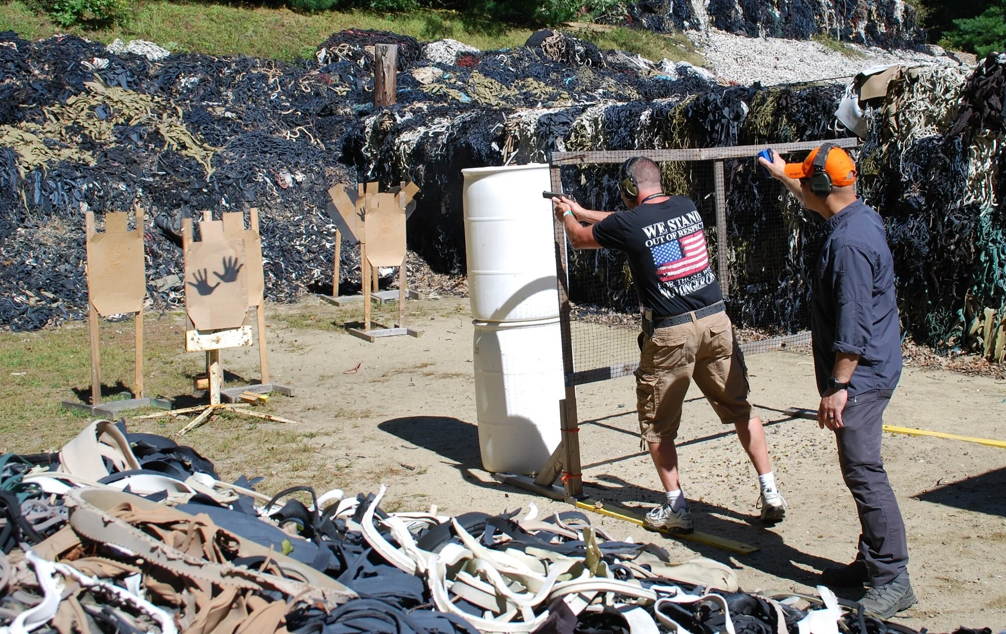 Two men at a shooting range with silhouettes of hands on targets, shooting at clay pigeons with a backdrop of black and green rubber waste.