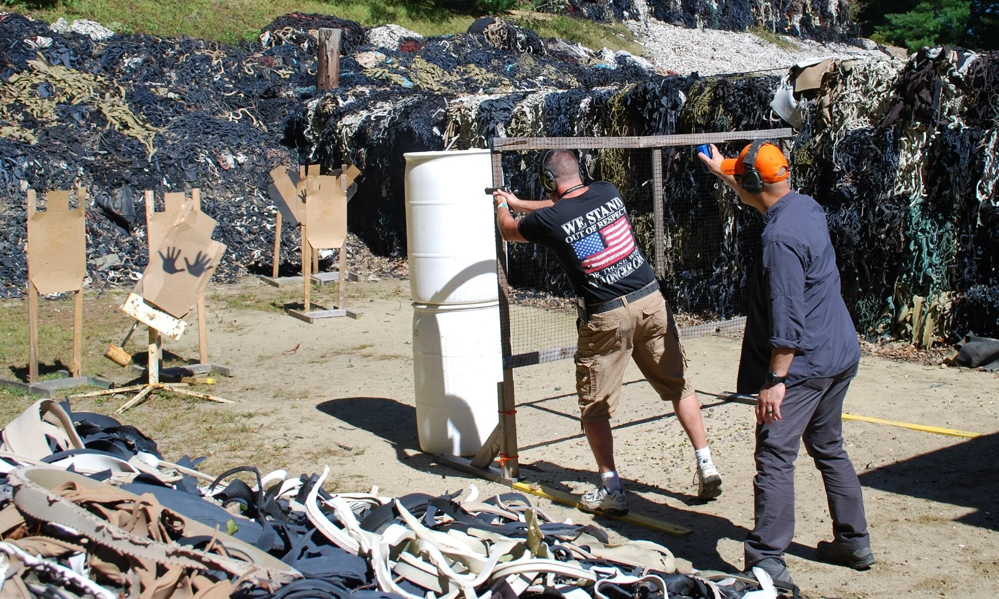 Two men practicing shooting at outdoor firing range with cardboard targets and rubber material in background.