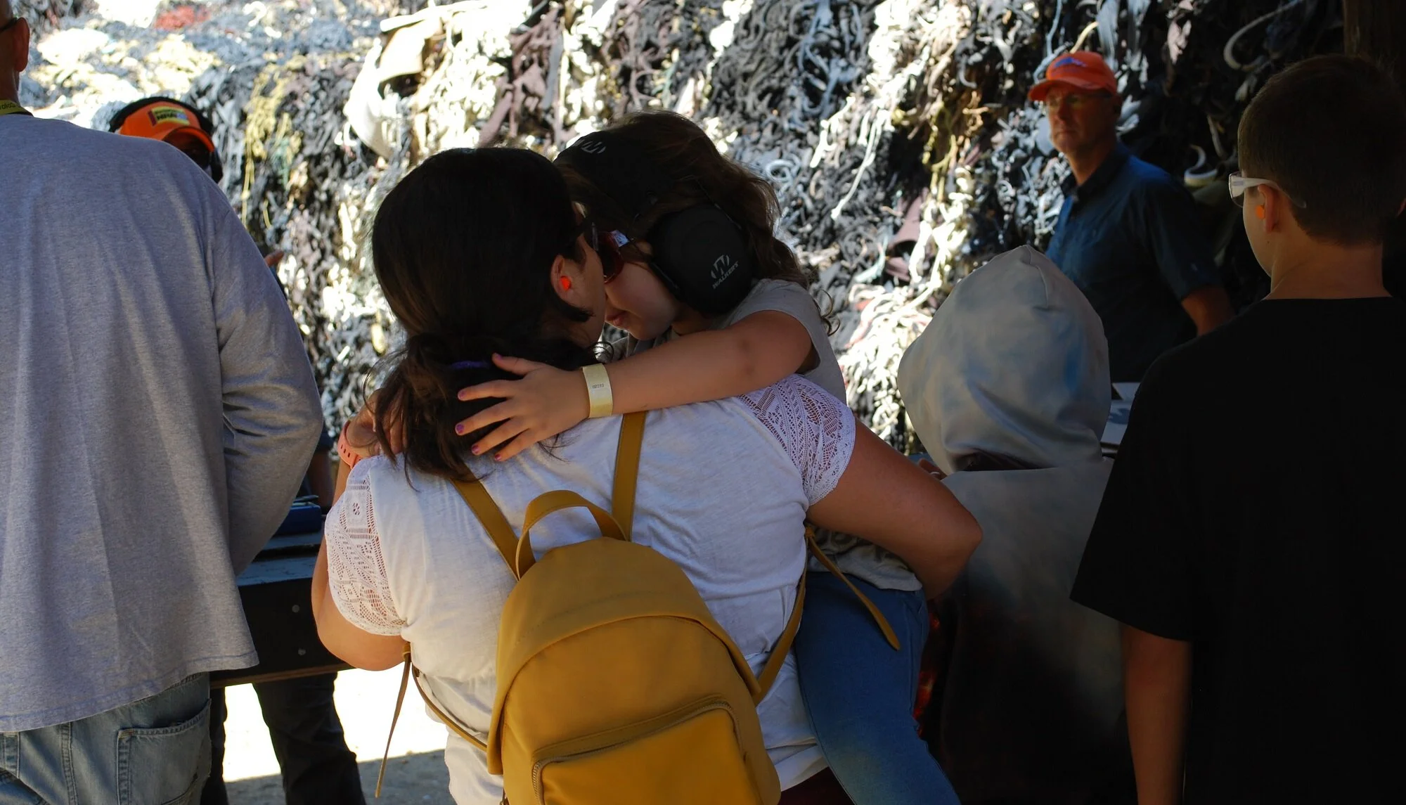 Two young girls embrace each other at an outdoor event, surrounded by other people, with a background of a rocky wall.