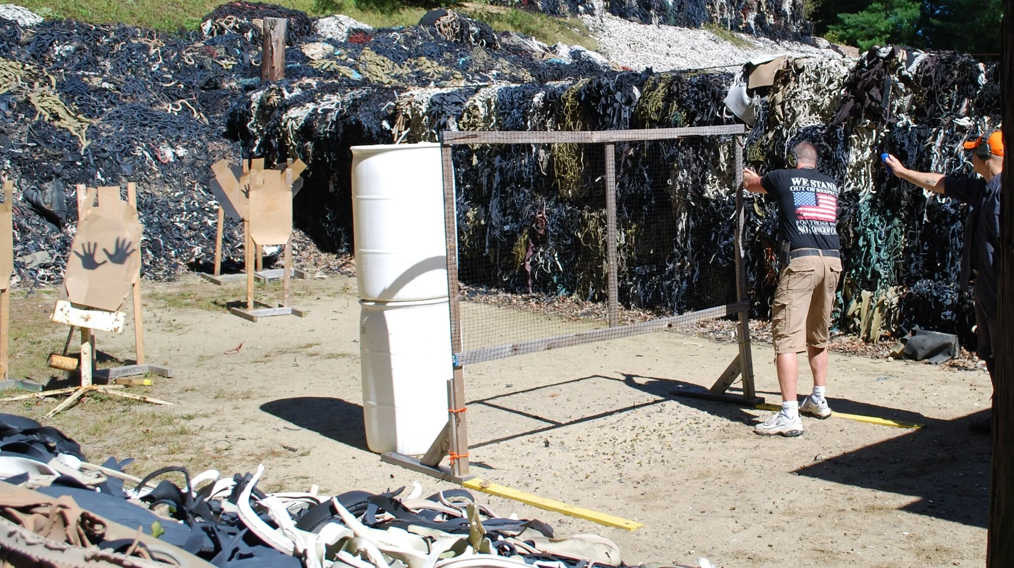People shooting at paper targets with handprints on a dirt outdoor range, with a large pile of shredded rubber and tires in the background.