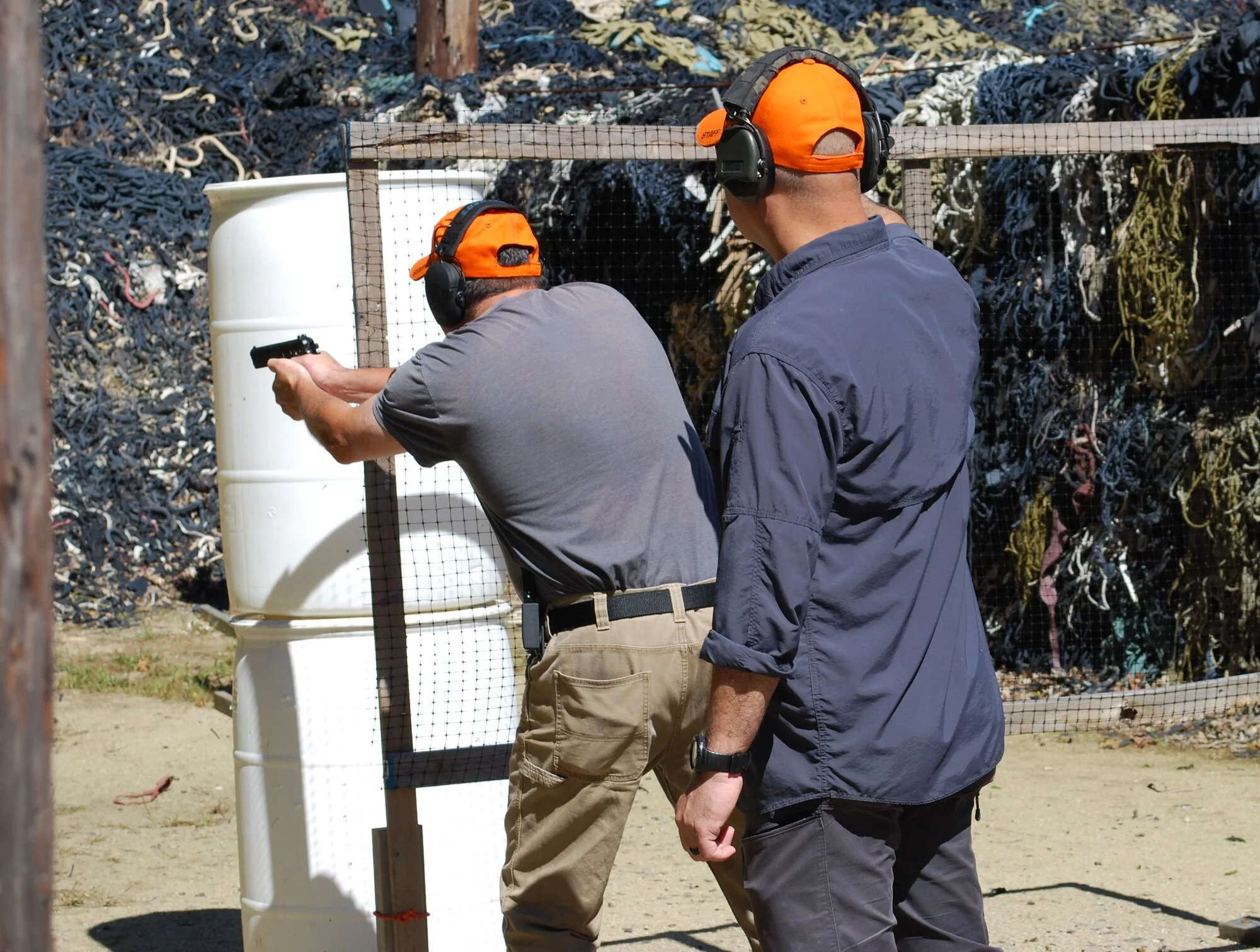 Two men with orange caps and ear protection practicing shooting at a gun range. One man is aiming a handgun, while the other observes.