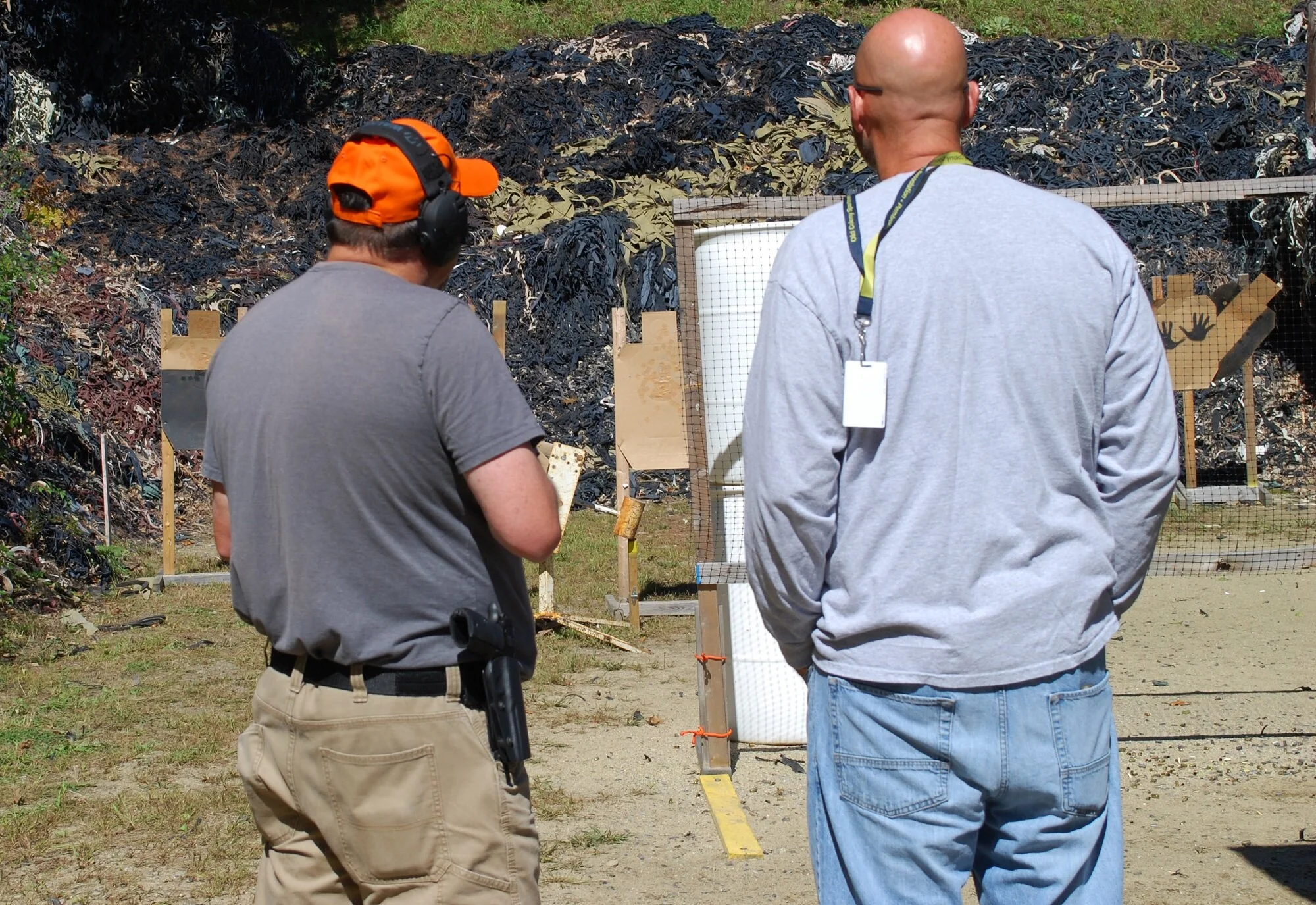 Two men at an outdoor shooting range, one wearing an orange hat and headphones, the other bald, overlooking targets and a backdrop of rubber debris.