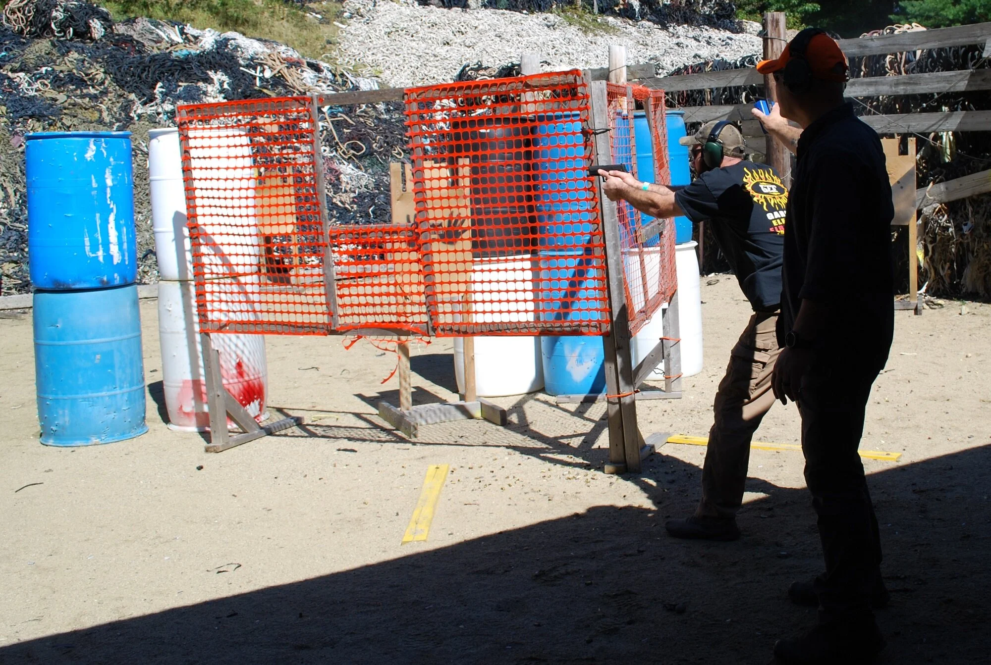 People shooting at an outdoor shooting range, with barrels and obstacles, in a sunny environment.