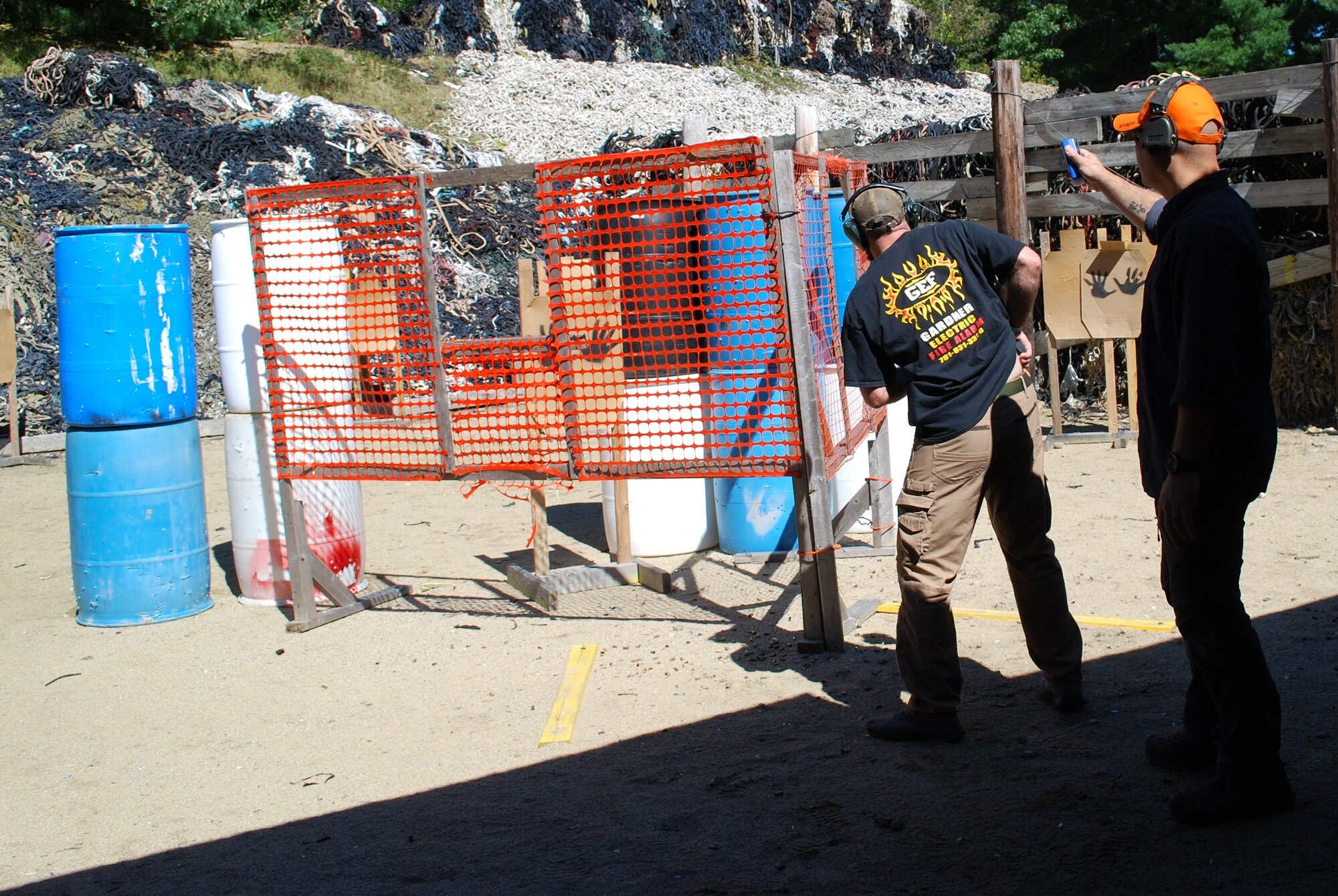 Two men at a shooting range with barrels and a target behind a fenced area, one with a timer, both wearing headphones.