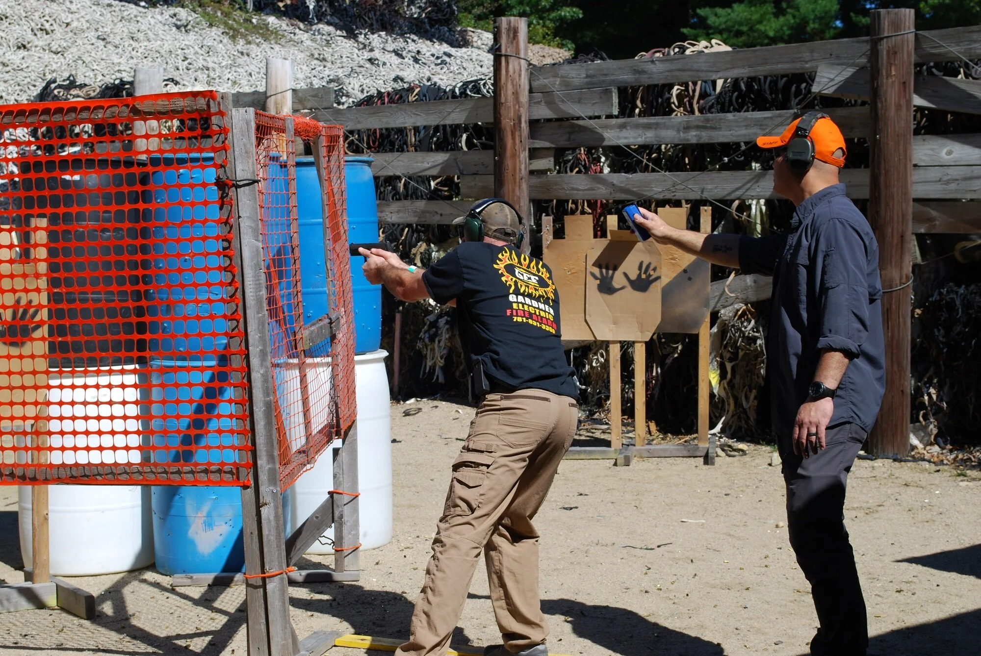 Two men at a shooting range, one aiming a firearm and the other with a timer, blue barrels and a wooden fence in the background.