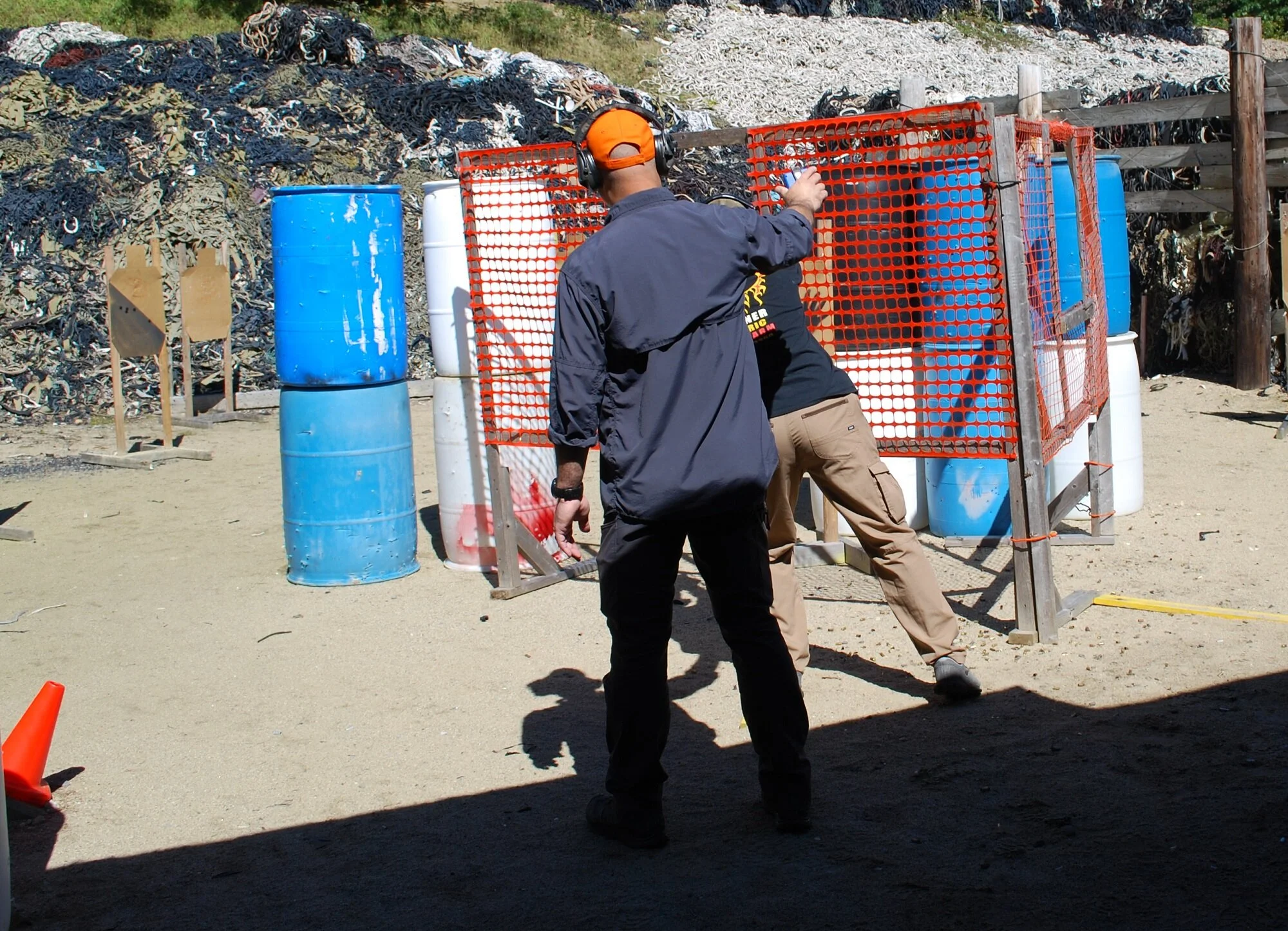A man wearing a black jacket, khaki pants, and an orange cap is practicing shooting at a shooting range with a person behind a safety barrier, with blue and white barrels and a dirt ground.