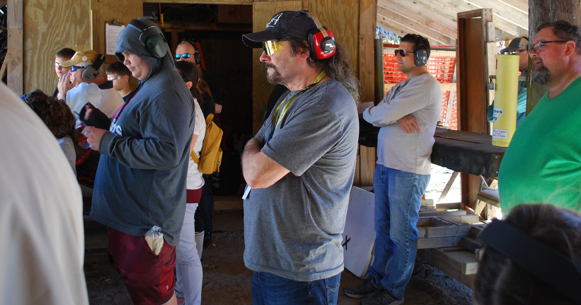 Group of people at a shooting range, wearing hearing protection and safety glasses, standing inside a wooden structure, some focused on something off-camera.