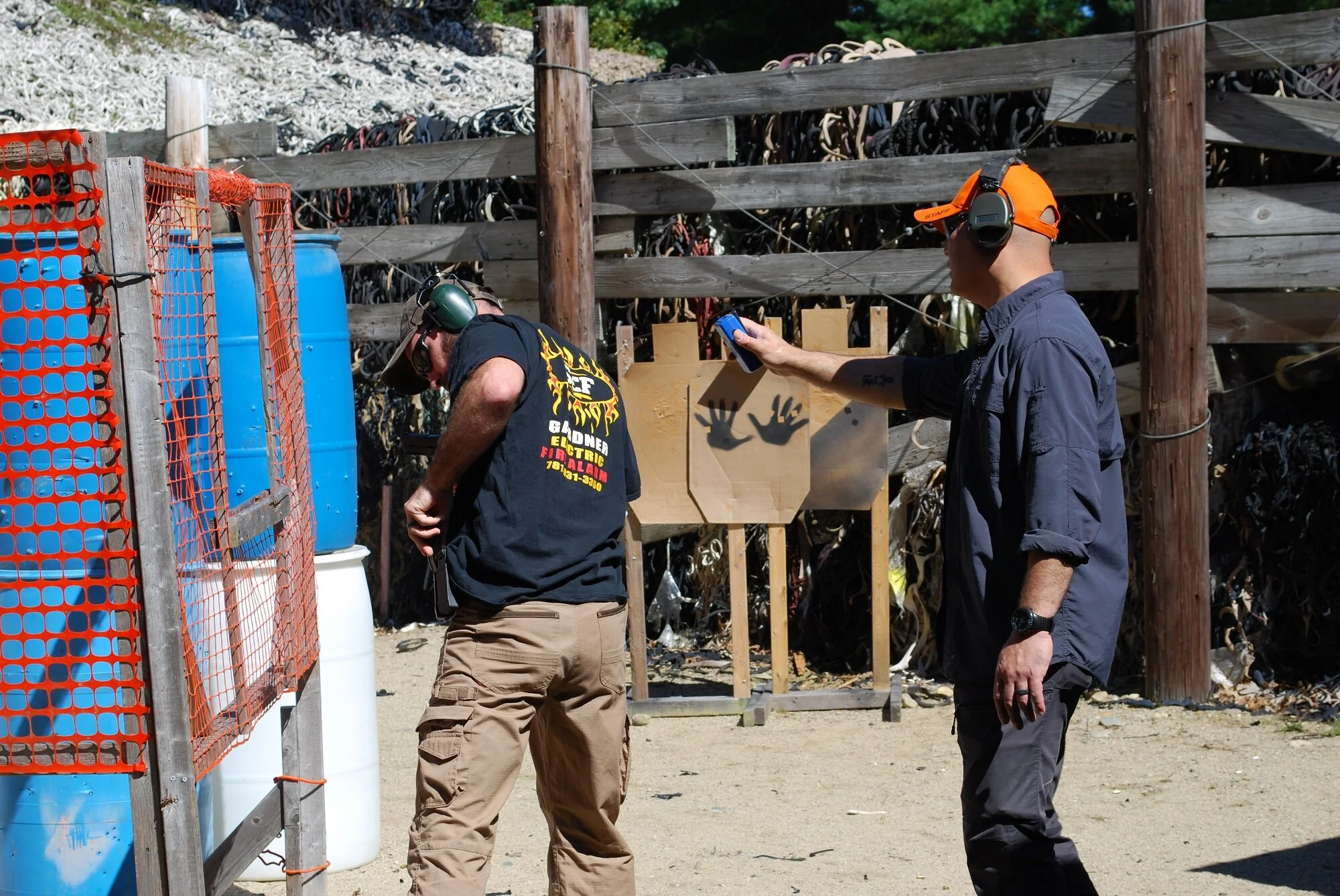Two men at a shooting range wearing ear protection, with one man aiming a handgun and the other a timer. There are blue barrels, a wooden fence, and a target with handprints in the background.