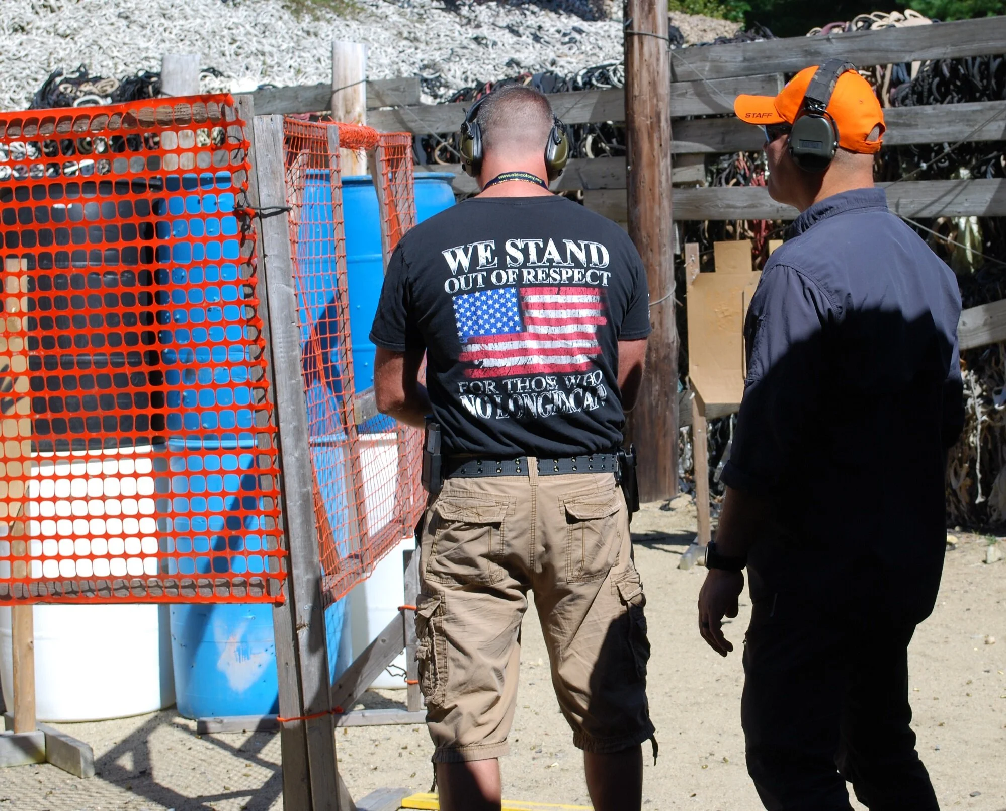 Two men at a shooting range, both wearing protective ear gear, with one wearing a shirt that says 'We Stand Out Of Respect For Those Who No Longer Can.' They are standing near a wooden fence with orange safety netting and large blue barrels and tires