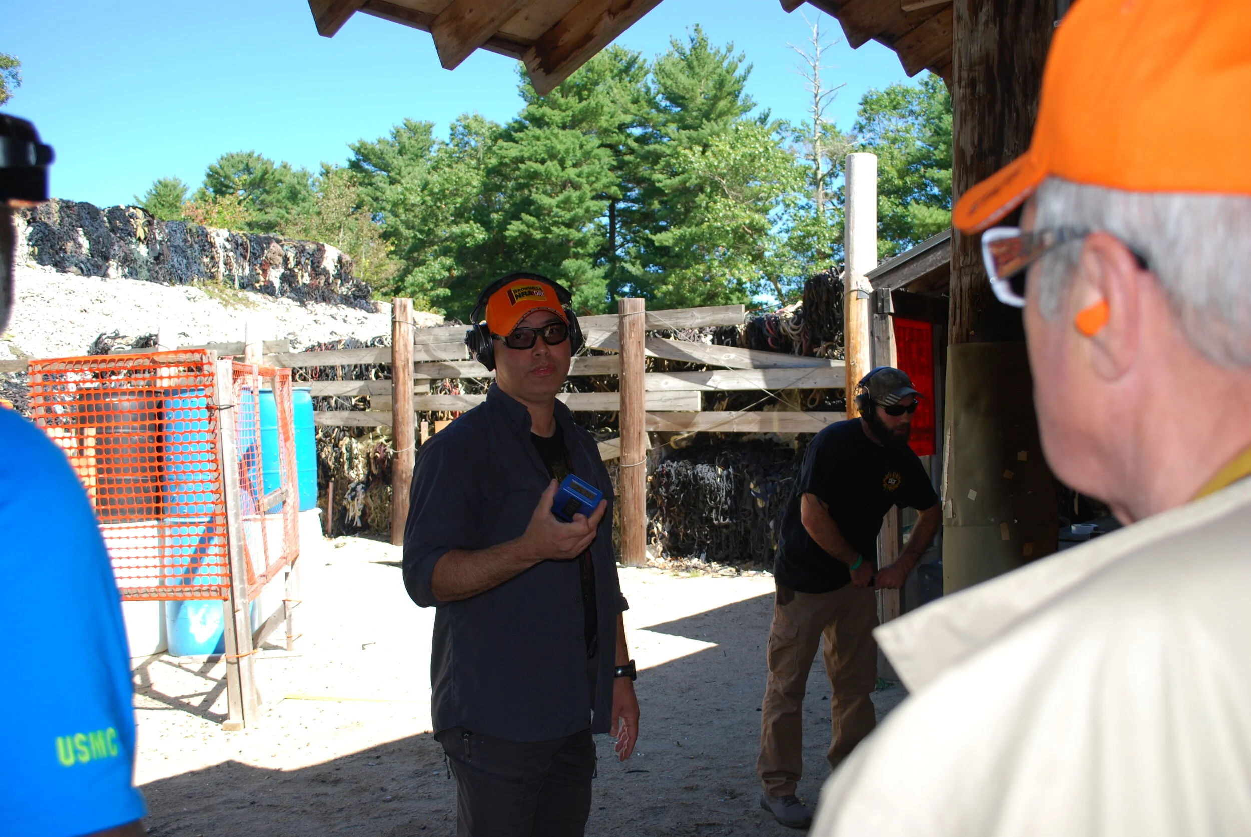 Four people at a shooting range, wearing hearing protection and safety glasses. The person in the foreground is an elderly man with white hair, wearing an orange cap and ear protection. The person in the middle is a man holding a box, wearing a black