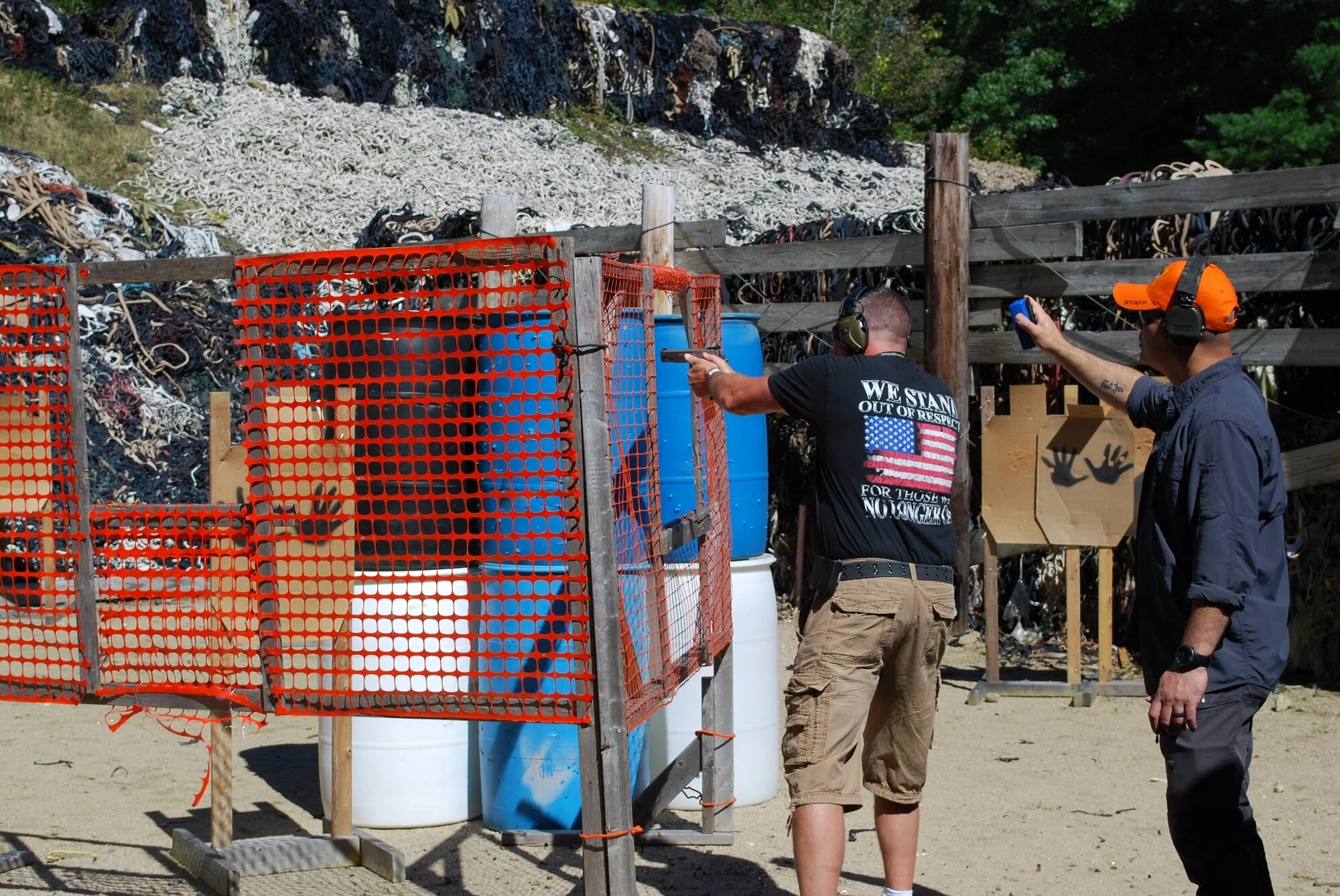 Two men at a shooting range aiming their firearms at paper targets, with large barrels and a wall of trash in the background.