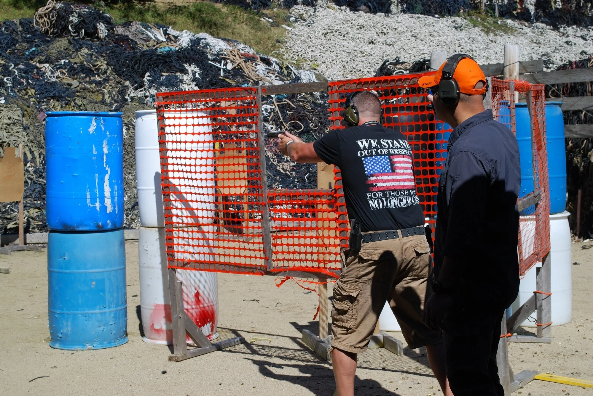 Two men at an outdoor shooting range, with barrels, orange safety barricades, and a background of debris and natural terrain.