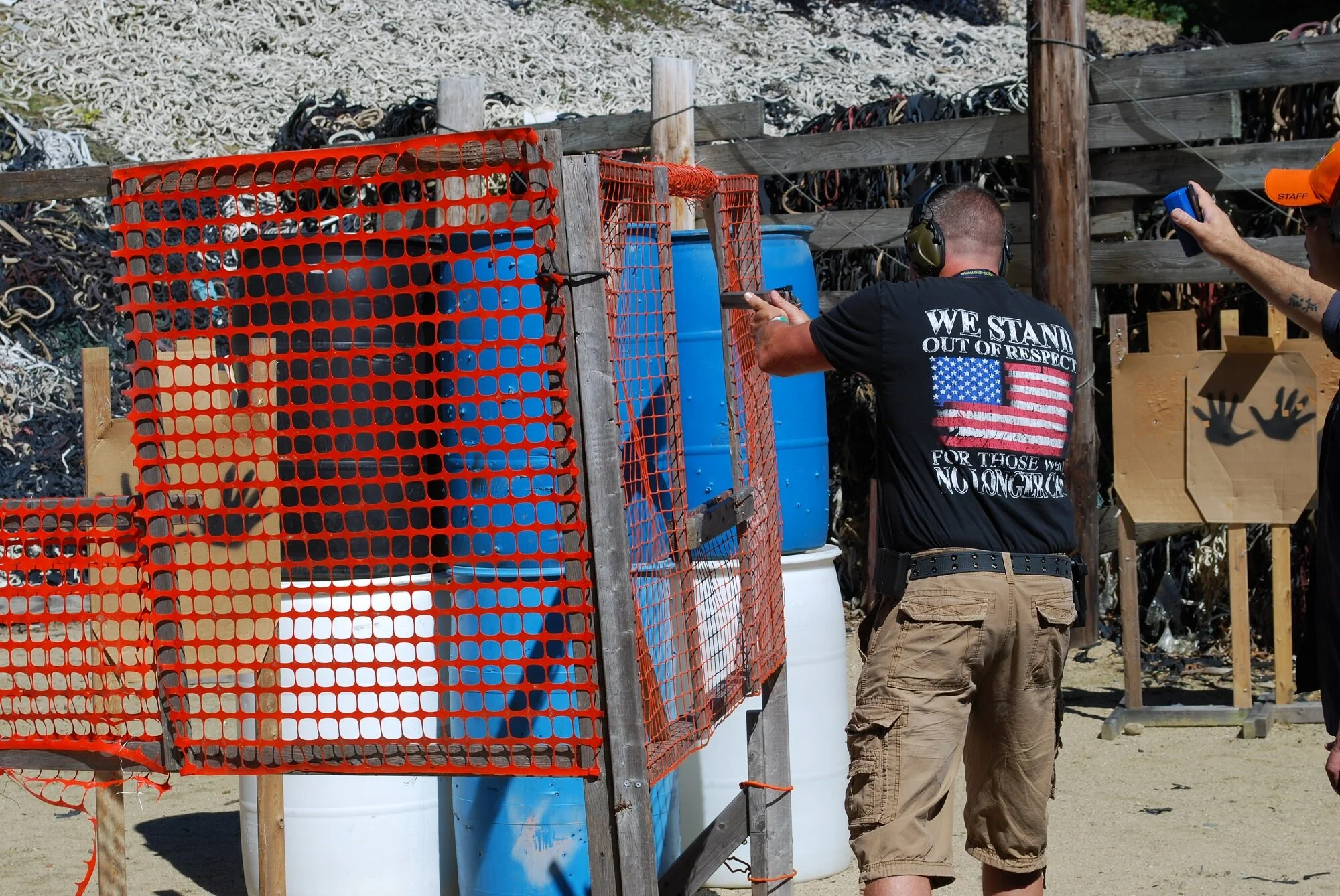 A person aiming a firearm at a shooting range with targets and safety barriers, wearing ear protection and a T-shirt that reads "We Stand Out of Respect For Those Who No Longer Can"