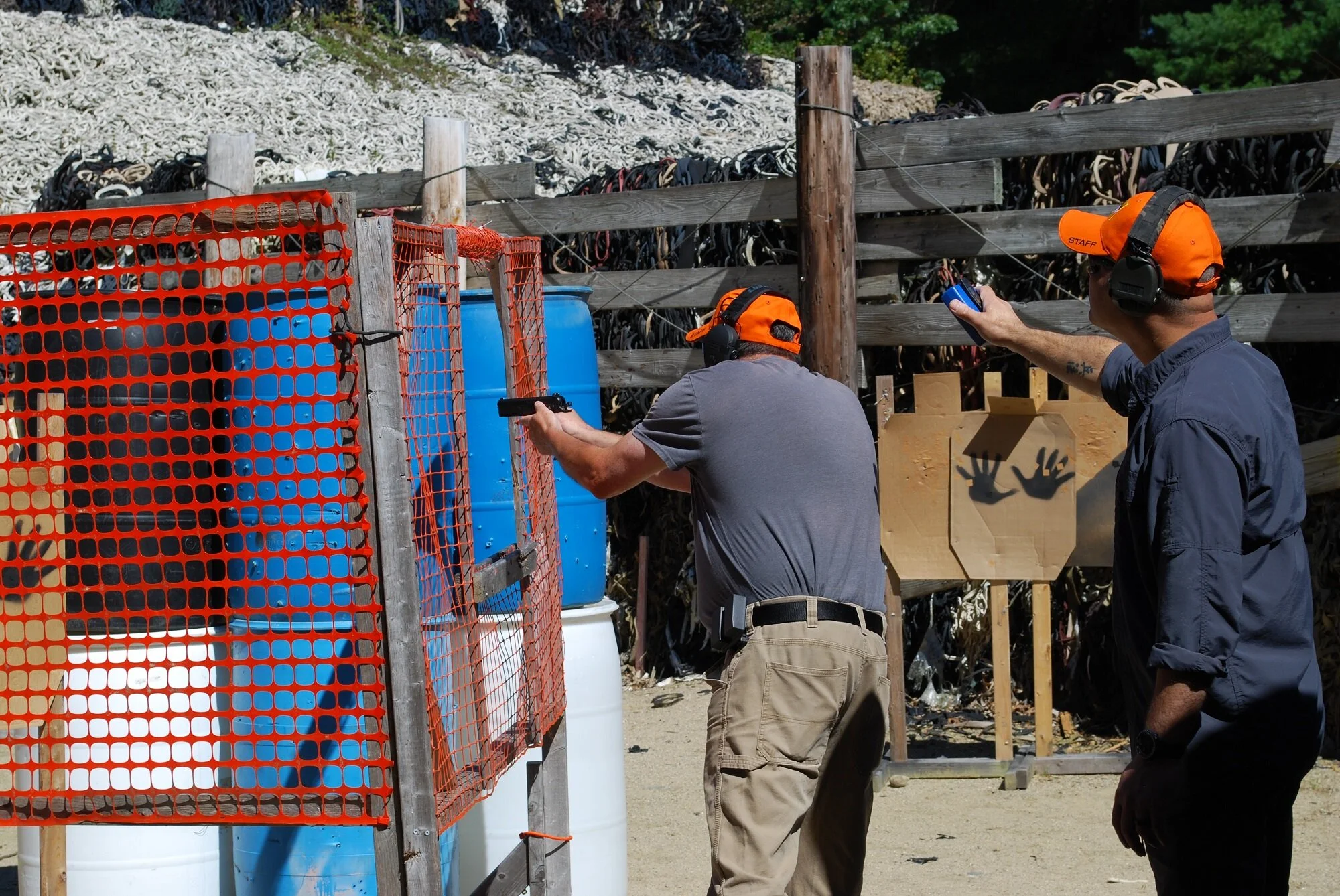 Two men at a shooting range, one aiming a handgun while the other appears to be giving instructions, both wearing orange caps and hearing protection, with targets and barrels in the background.