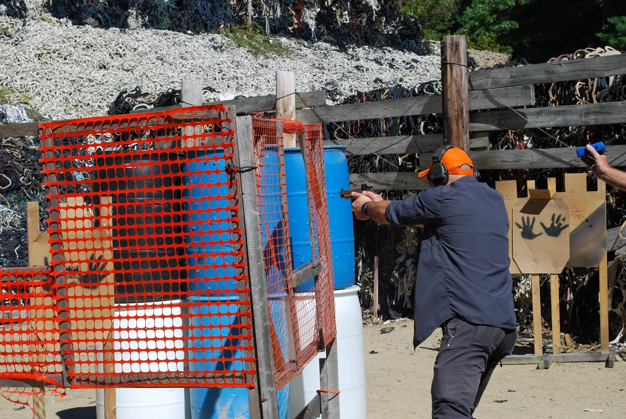 A man at an outdoor shooting range aiming a pistol at a target. The man is wearing ear protection and an orange cap. There are orange safety barriers and barrels around. In the background, there's a pile of used rubber and a wooden fence.