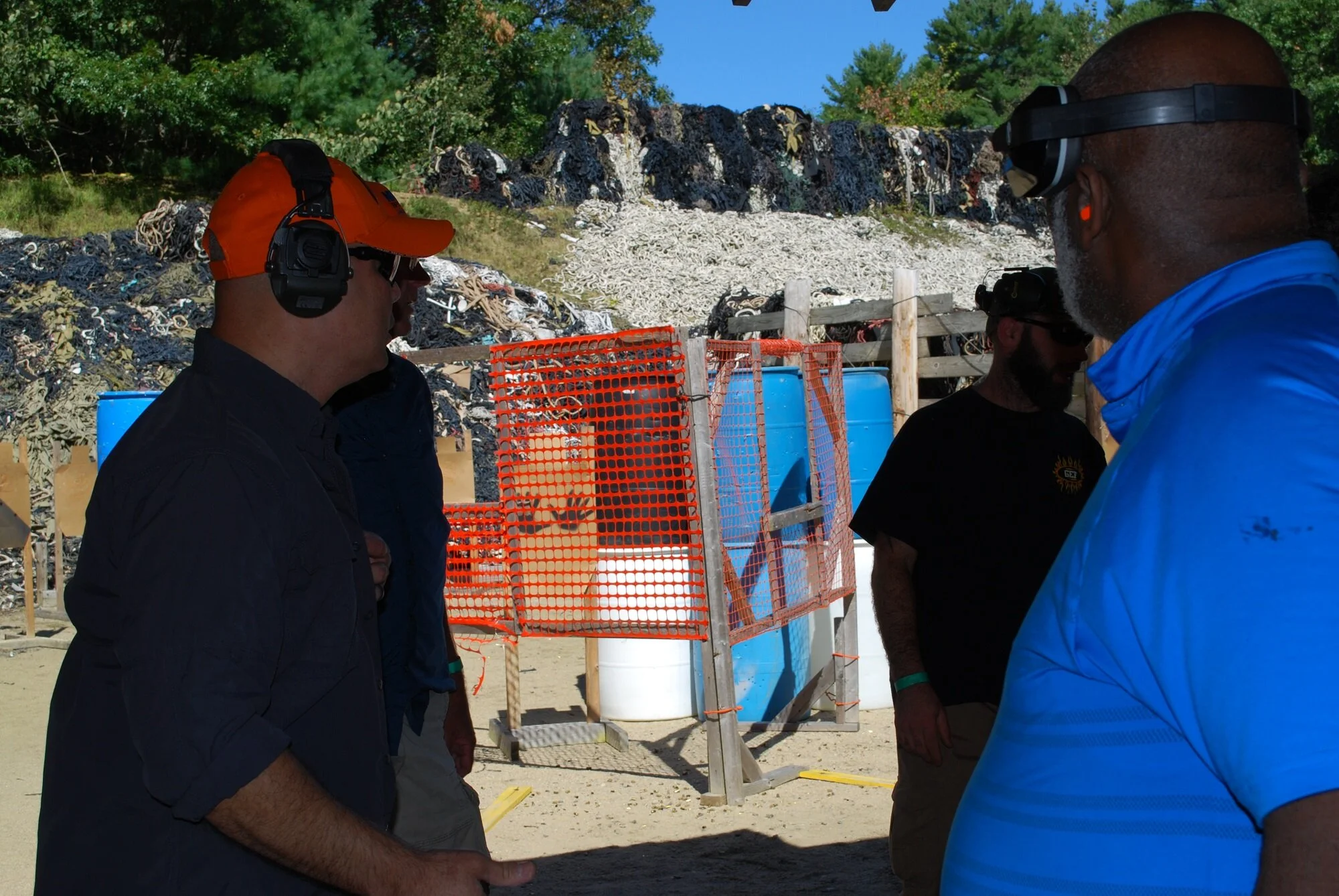 Group of men wearing safety vests and headphones at an outdoor shooting range, with targets and barrels in the background.