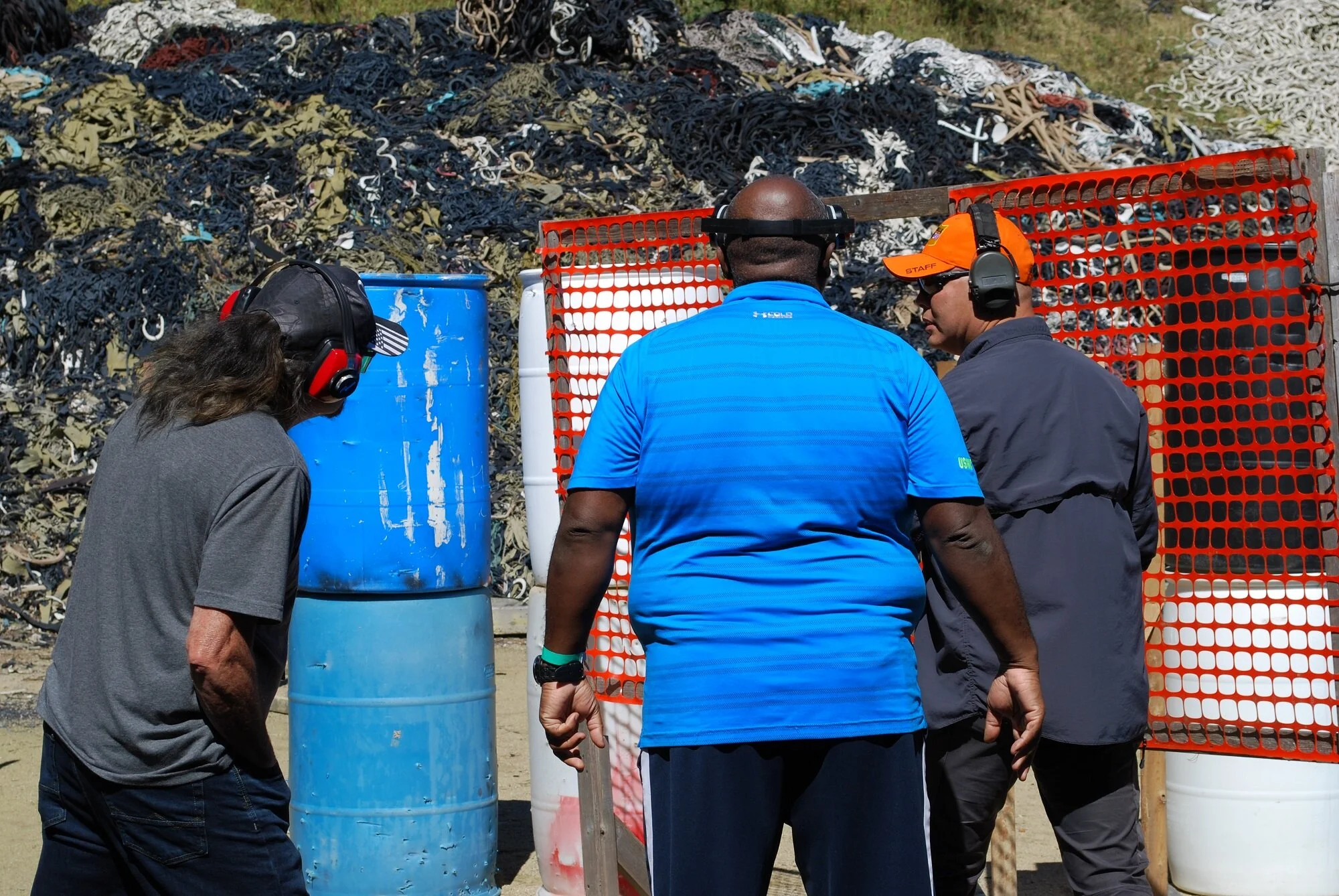 Three people participating in a shooting activity at a gun range, wearing protective ear and eye gear, with one person aiming at a target behind orange safety barriers, and a backdrop of debris.