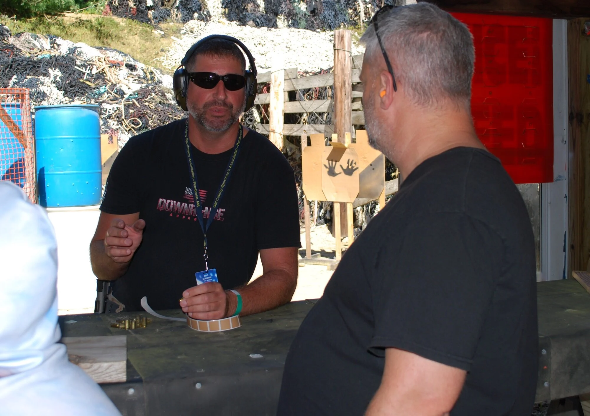 Two men engaged in conversation at an outdoor shooting range. One man wears sunglasses, hearing protection, and a black t-shirt, while the other has gray hair, a black shirt, and sunglasses resting on his head. Background includes a pile of scrap, a 