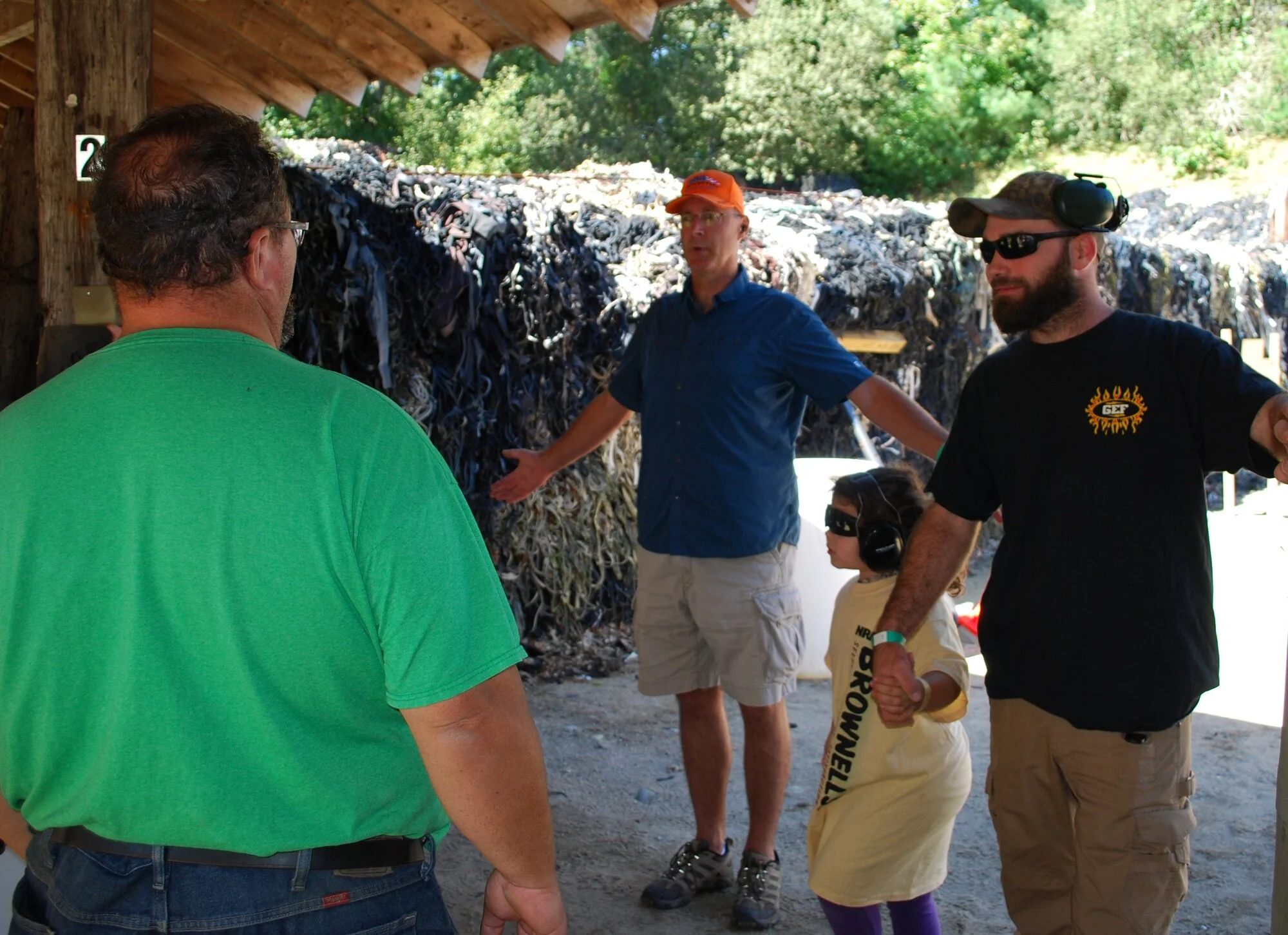 Group of people at an outdoor shooting range, wearing ear protection, with a large pile of rubber in the background.