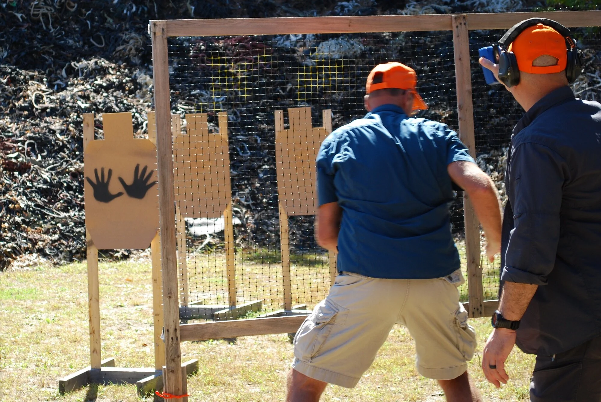 Two men practicing combat shooting at an outdoor shooting range with cardboard targets and safety gear.