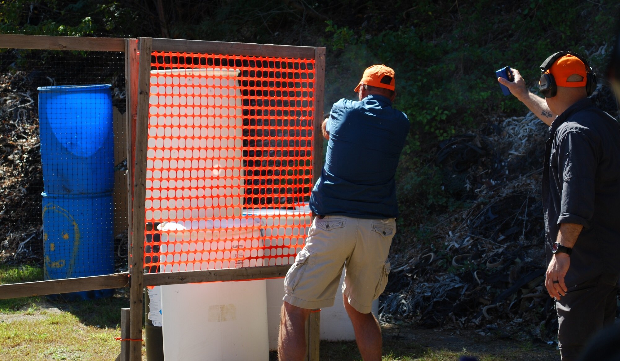 Two men shooting at a paper target behind a temporary barricade made of wood and orange plastic mesh, outdoors near a pile of scrap rubber and foliage.