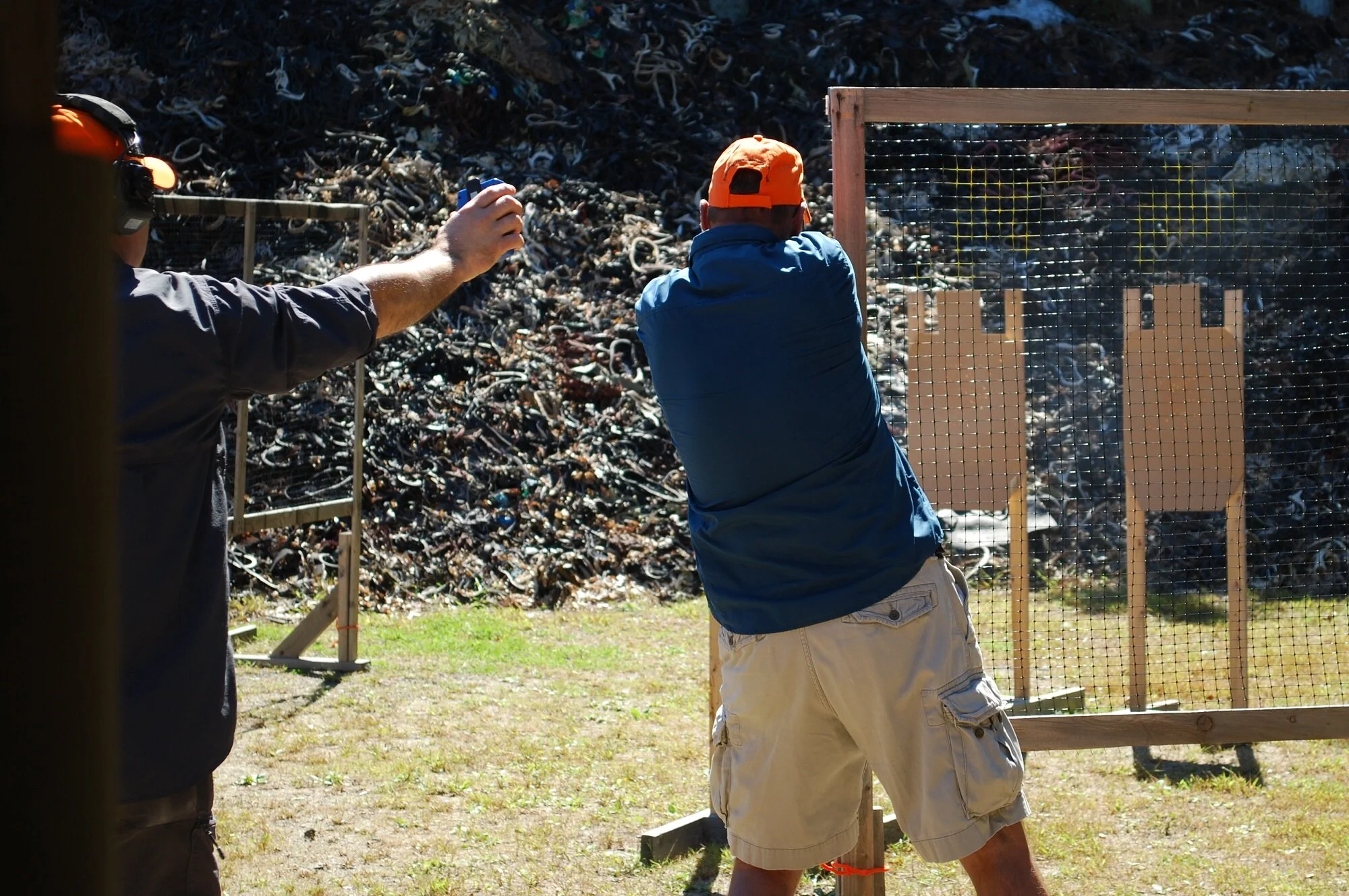 Two men practicing shooting at an outdoor gun range with targets and large pile of scrap rubber in the background.