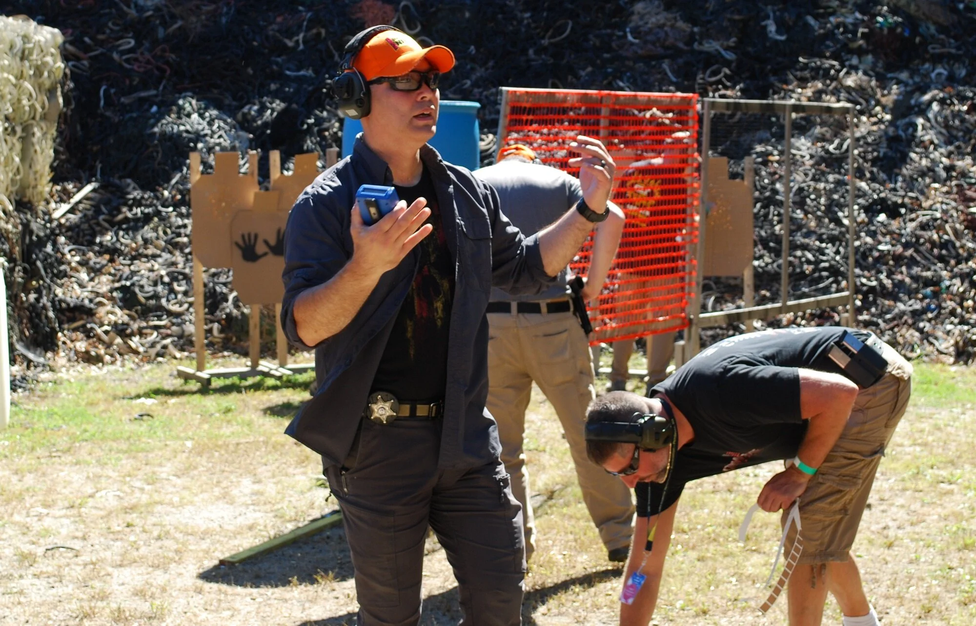 Two men in black T-shirts and khaki shorts, wearing headphones and sunglasses, are at an outdoor shooting range. One man in a dark shirt is standing and talking, while the other is bent over, reviewing something on the ground. The background has a he