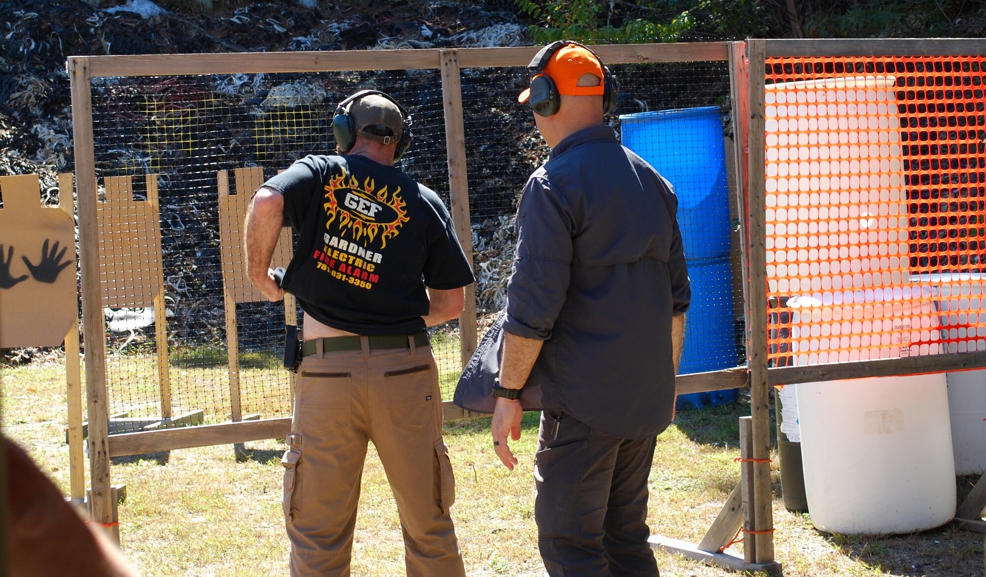 Two men at a shooting range preparing, with noise-canceling earmuffs, in front of a makeshift target with orange mesh and blue barrels.