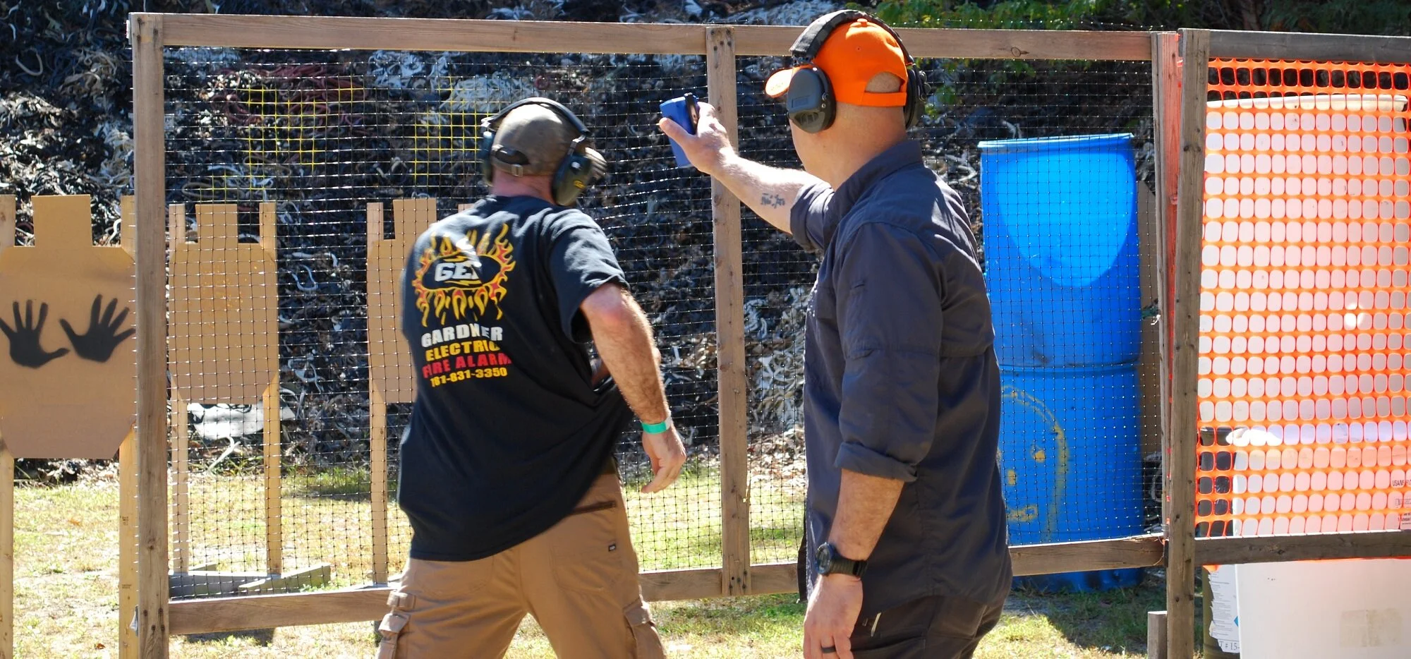 Two men at an outdoor shooting range, one instructing the other. They are wearing ear protection and casual clothing. The background consists of various colorful target boards and a wooden fence.