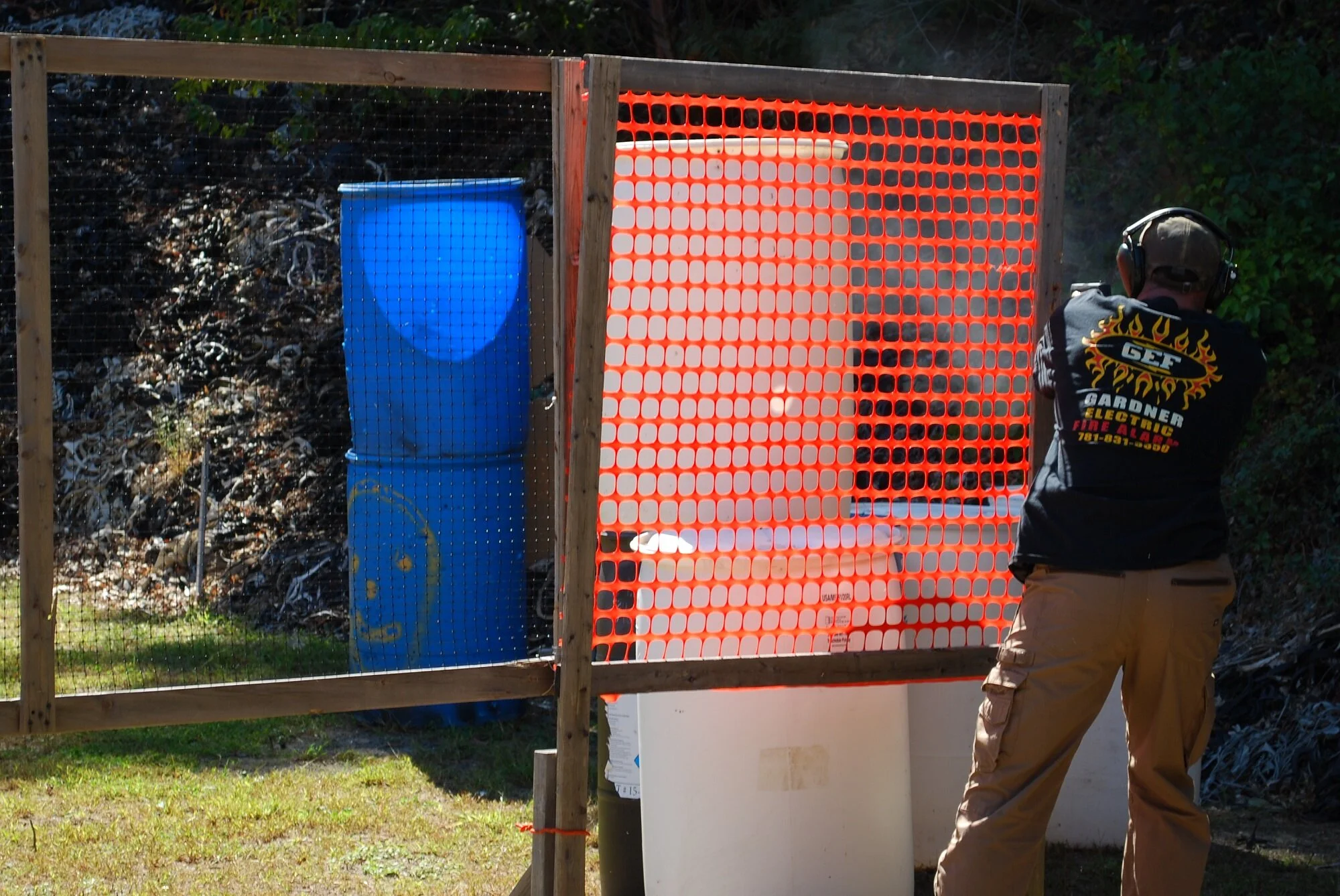 A man wearing a black shirt with fire graphics and tan cargo pants, and a headset, shooting at an outdoor shooting range. He is standing behind a safety barrier made of wooden frames and orange safety netting.
