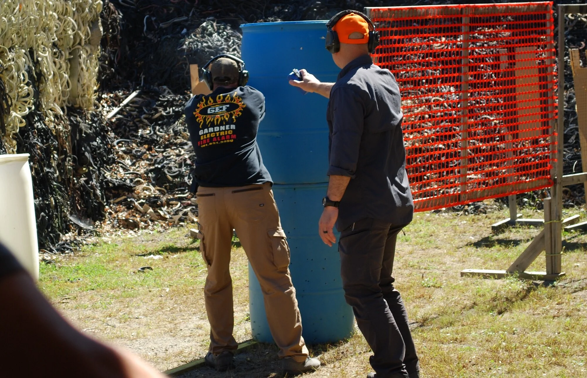 Two men wearing protective ear gear are at a shooting range, aiming a rifle at a target. One man is dressed in a black shirt with a flame logo and tan pants, while the other wears a dark blue shirt and dark pants.